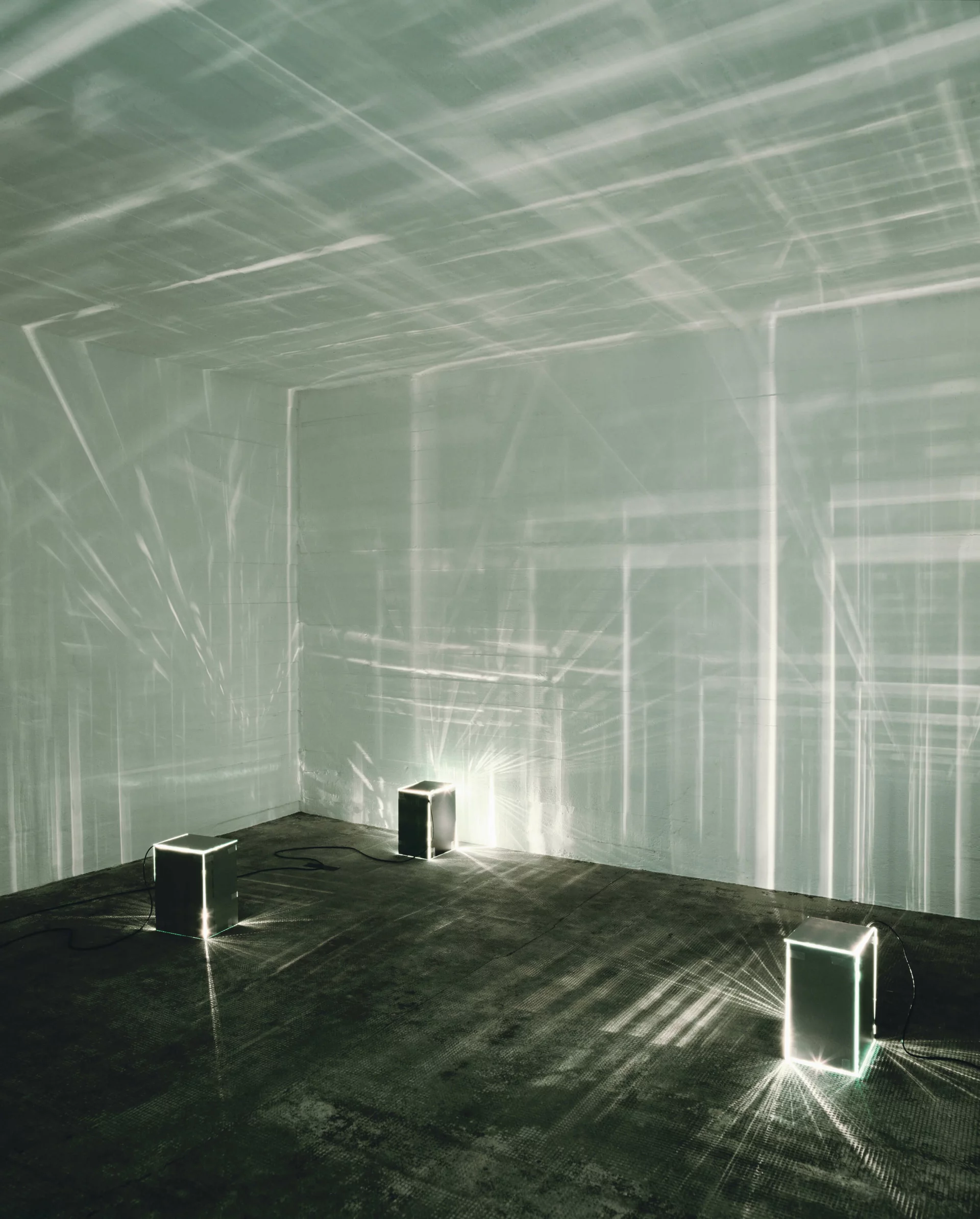 Vier leuchtende Glaskuben auf Boden werfen Lichtreflexionen an die Wände eines dunklen Raumes.