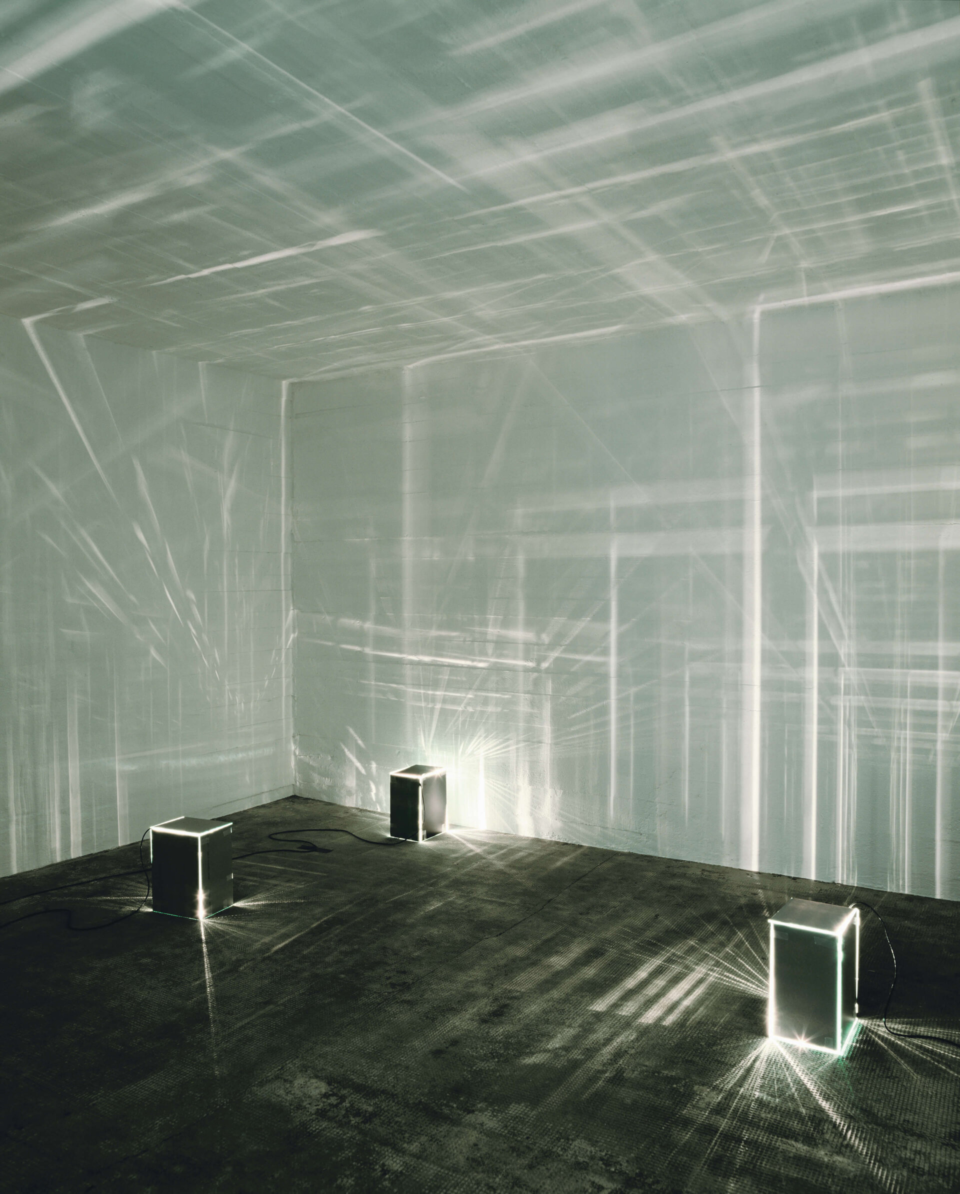 Vier leuchtende Glaskuben auf Boden werfen Lichtreflexionen an die Wände eines dunklen Raumes.