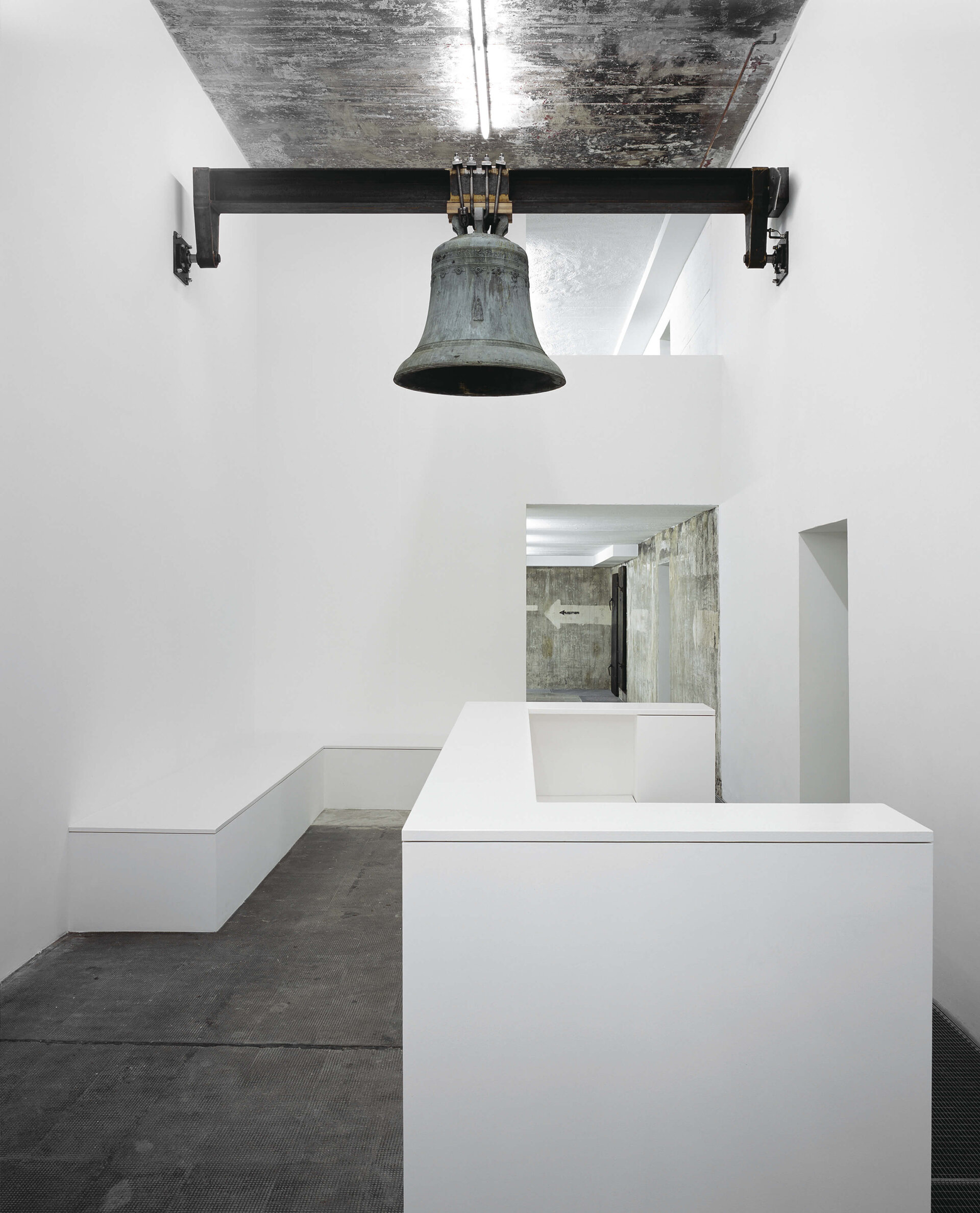 Große Glocke hängt von der Decke über einer minimalistischen weißen Empfangssituation in einer Ausstellungsarchitektur.