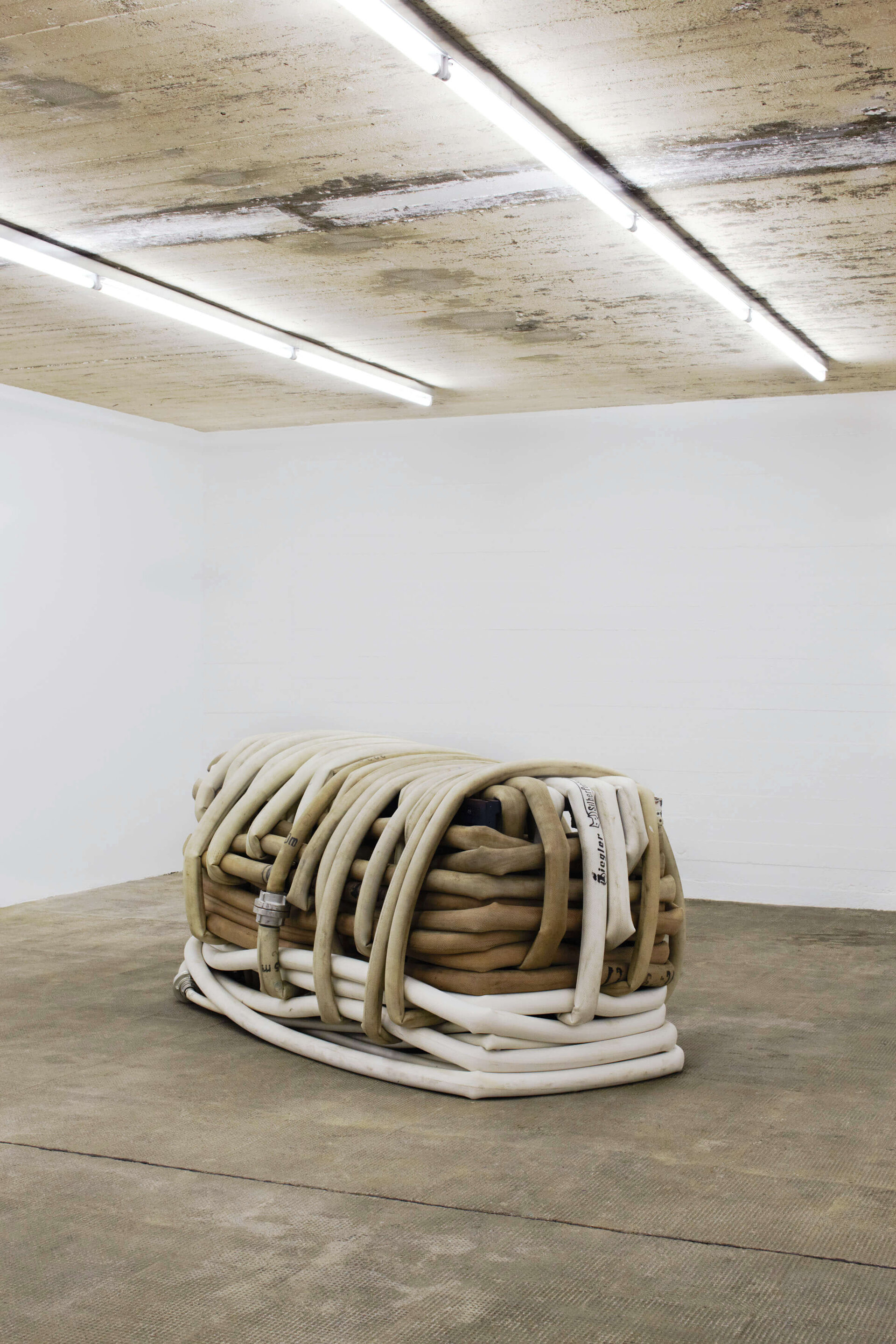Skulptur von Klara Lidén bestehend aus ineinander verschlungenen, gebrauchten Feuerwehrschläuchen in Beige- und Weißtönen, platziert zentral im Raum mit rohem Betonboden und weiß gestrichenen Wänden.