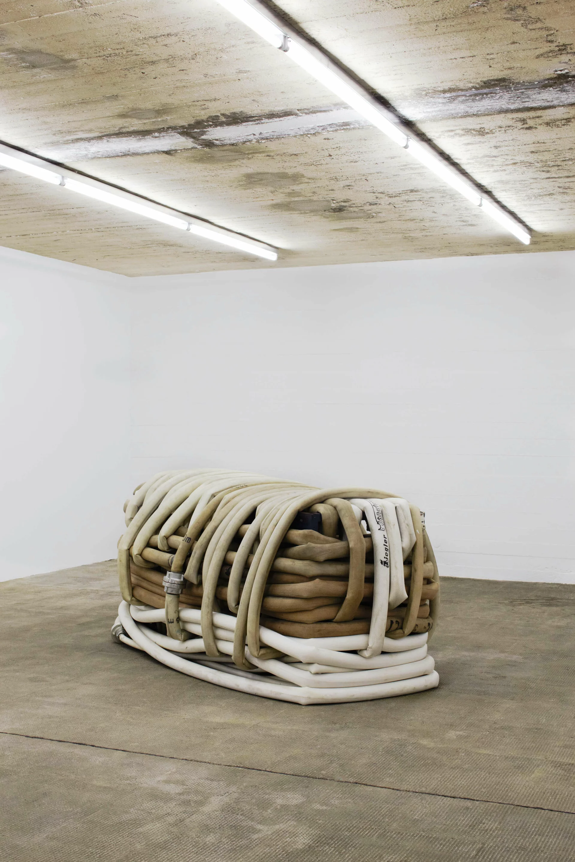 Skulptur von Klara Lidén bestehend aus ineinander verschlungenen, gebrauchten Feuerwehrschläuchen in Beige- und Weißtönen, platziert zentral im Raum mit rohem Betonboden und weiß gestrichenen Wänden.