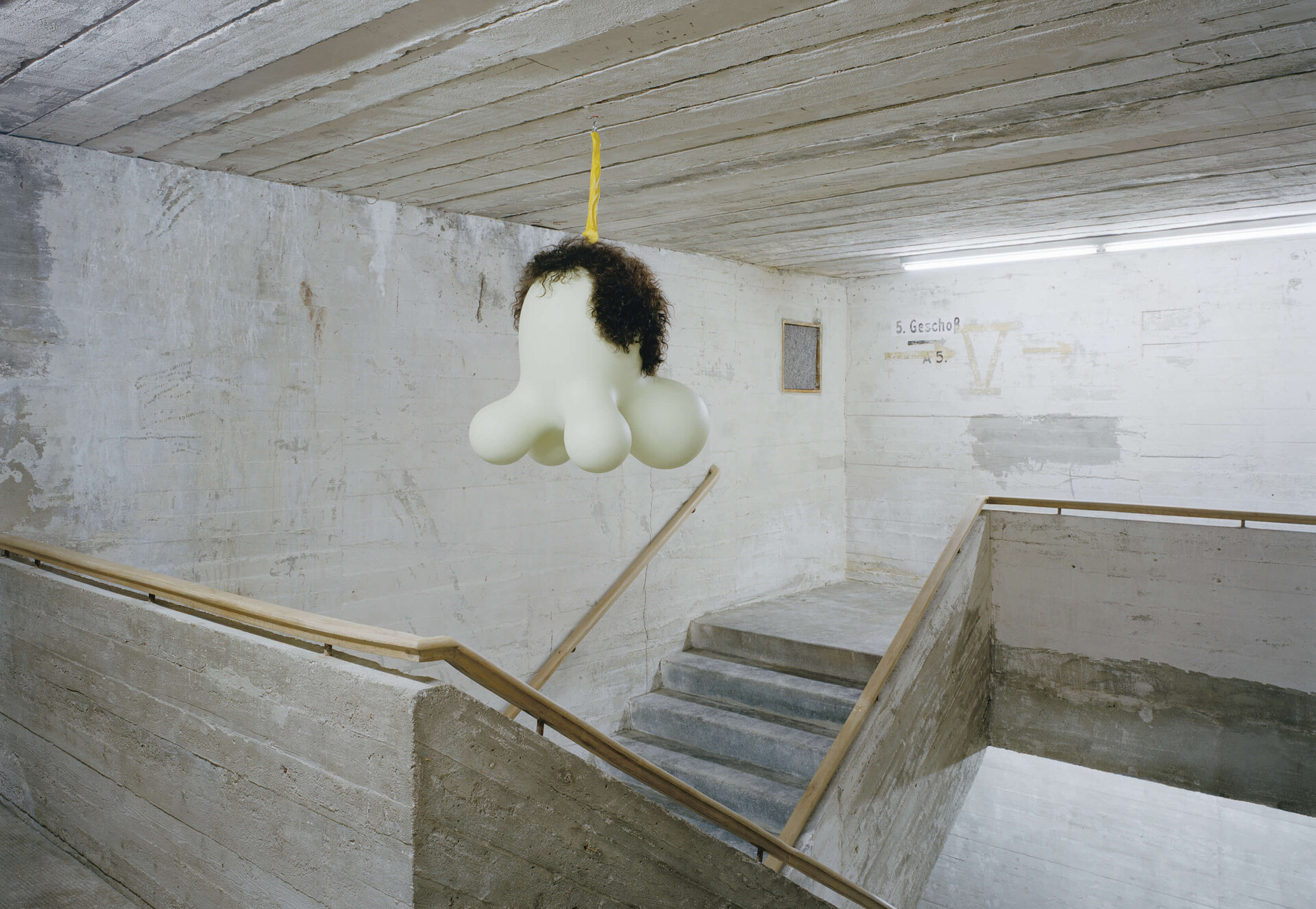 Organisch geformte Skulptur mit Perücke hängt an gelbem Band im Treppenhaus eines Betongebäudes.