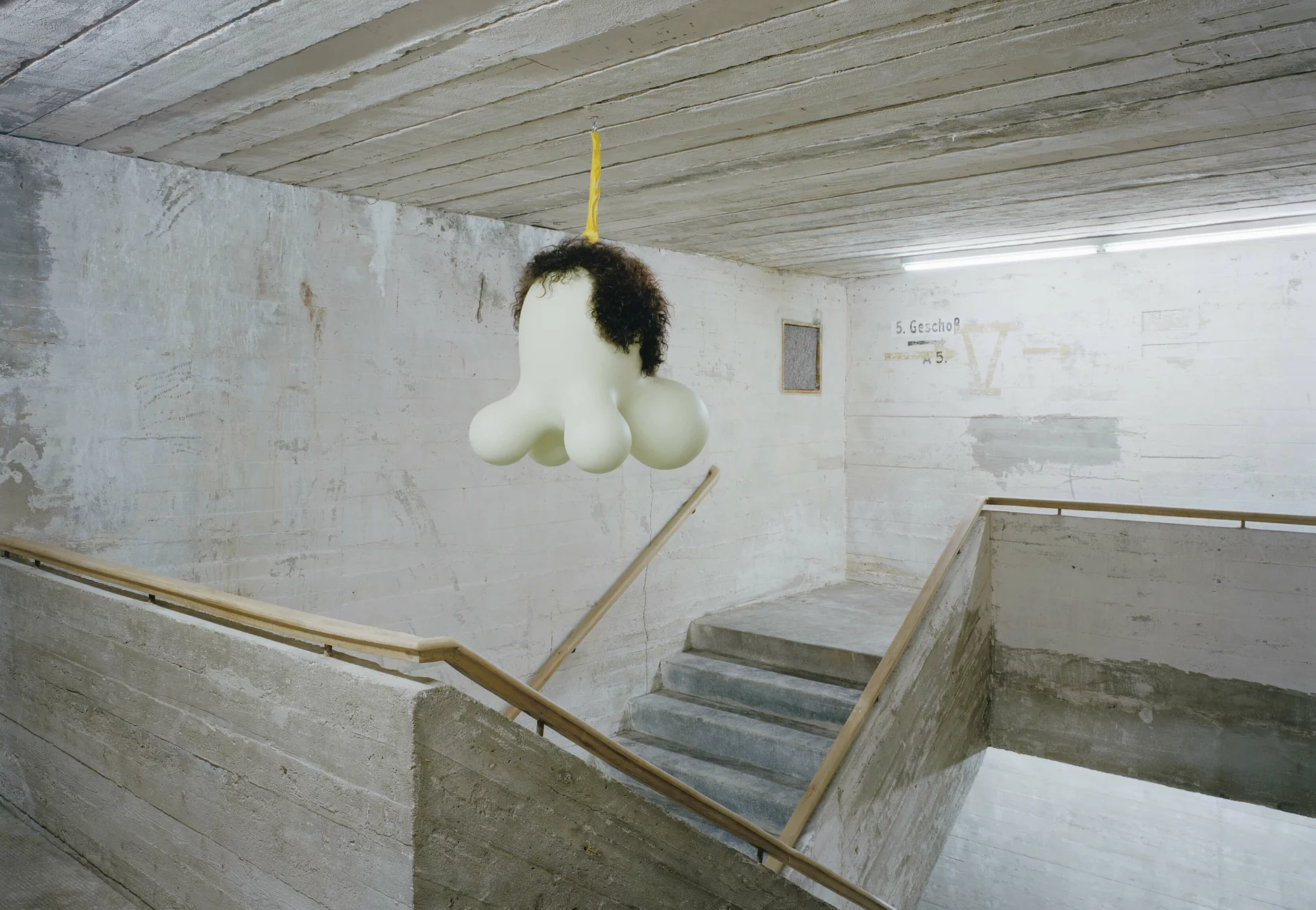 Organisch geformte Skulptur mit Perücke hängt an gelbem Band im Treppenhaus eines Betongebäudes.