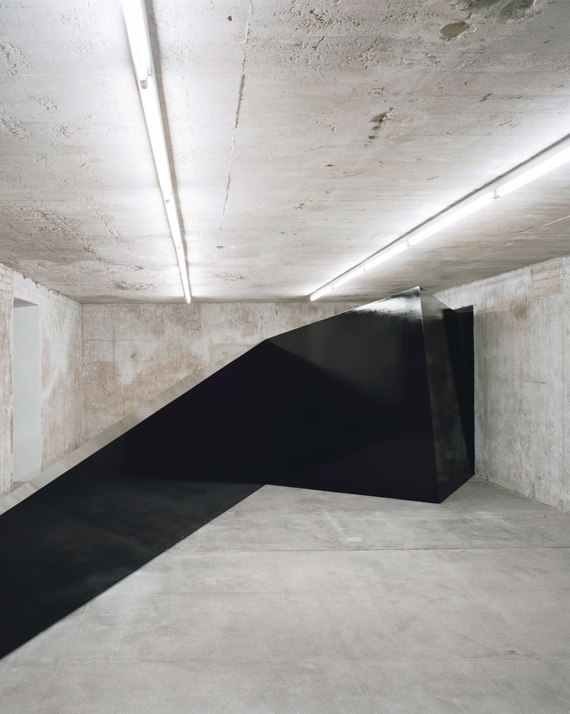 Schwarze, asymmetrische Metallskulptur schiebt sich diagonal durch den Raum eines Betonbunkers und kontrastiert mit der rauen Architektur.