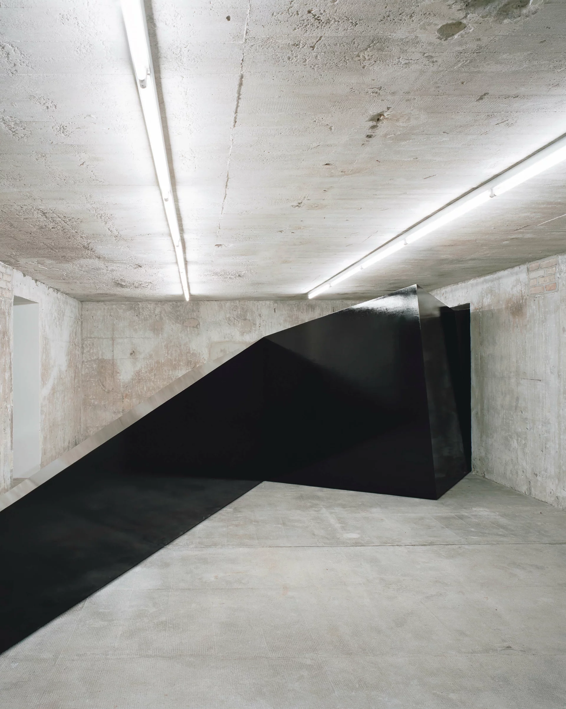 Schwarze, asymmetrische Metallskulptur schiebt sich diagonal durch den Raum eines Betonbunkers und kontrastiert mit der rauen Architektur.