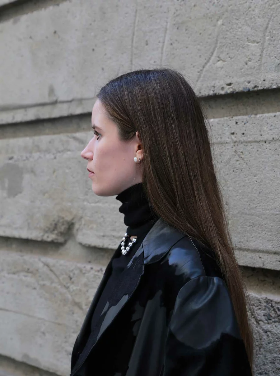 Porträt von Jil im Profil vor grauer Betonwand, mit schwarzem Outfit und Perlenkette, Teammitglied der Boros Collection.