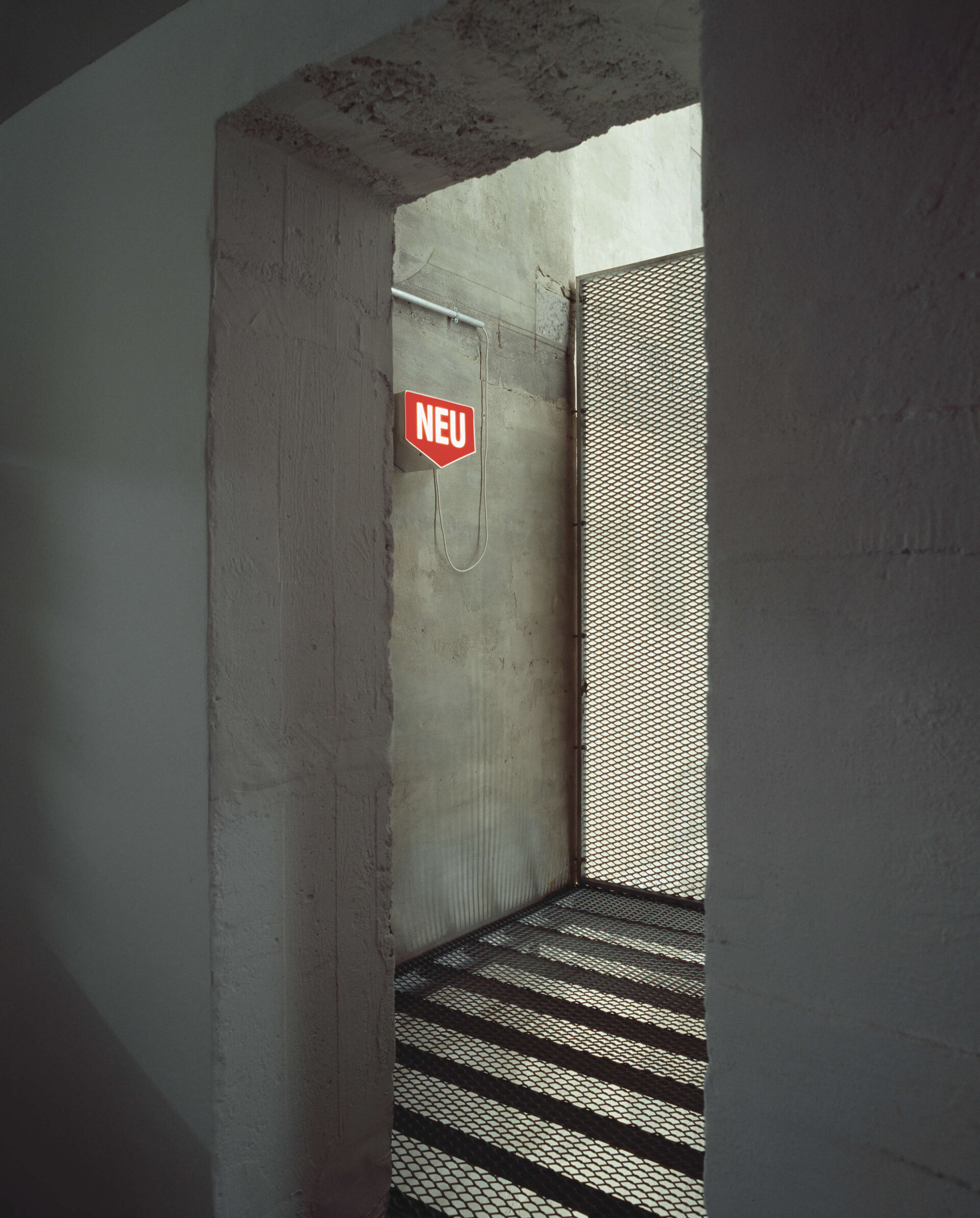 Leuchtschild mit „NEU“ in roter Schrift an Betonwand im Durchgang mit Gitterboden und Lichtspiel.
