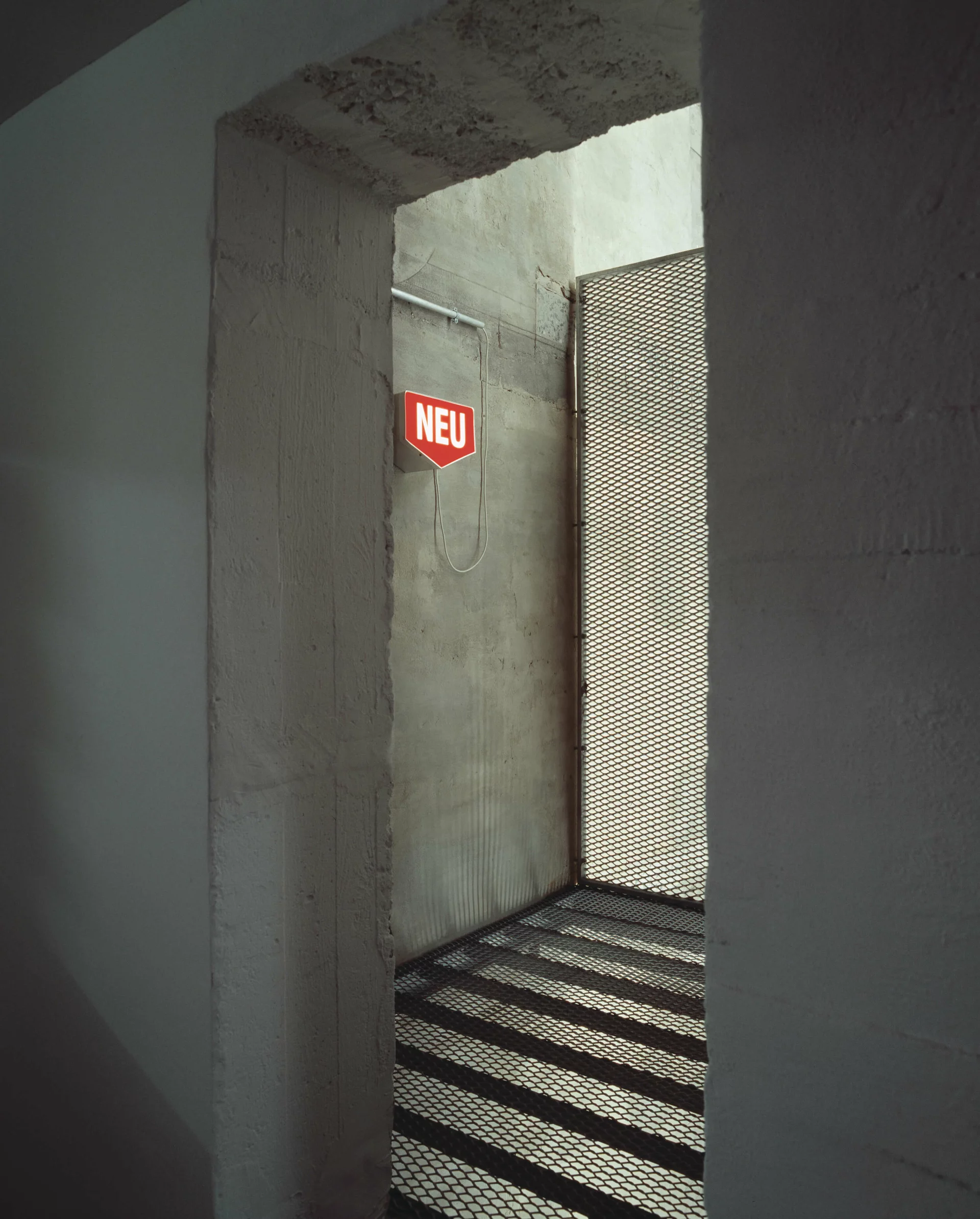 Leuchtschild mit „NEU“ in roter Schrift an Betonwand im Durchgang mit Gitterboden und Lichtspiel.