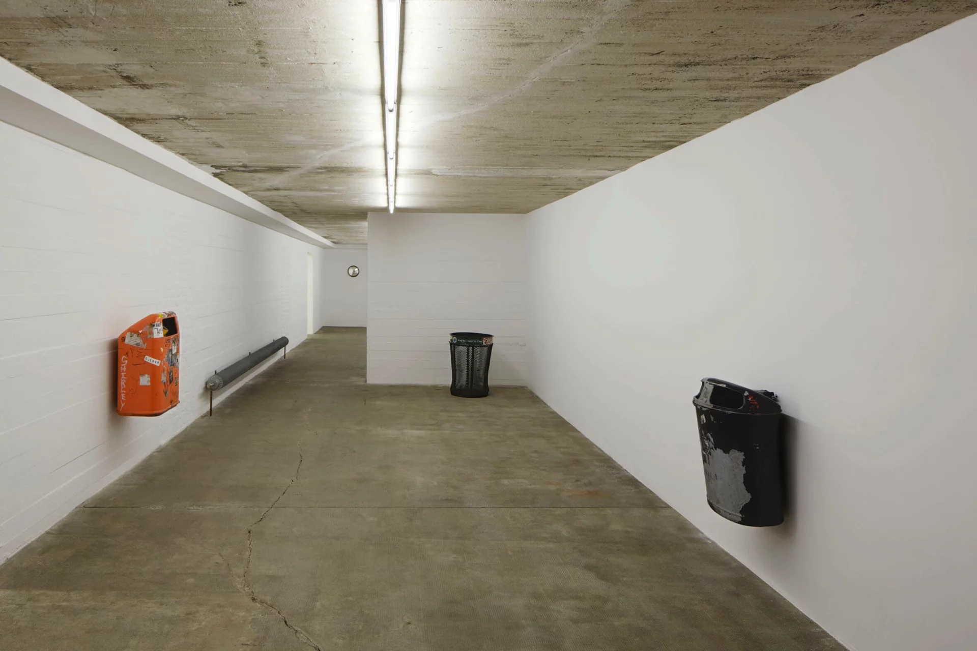 Drei gebrauchte Mülleimer unterschiedlicher Typen (orange, schwarz, grün) sind in einem weißen Ausstellungsflur montiert oder aufgestellt; das Setting erinnert an urbane Infrastruktur im musealen Kontext.