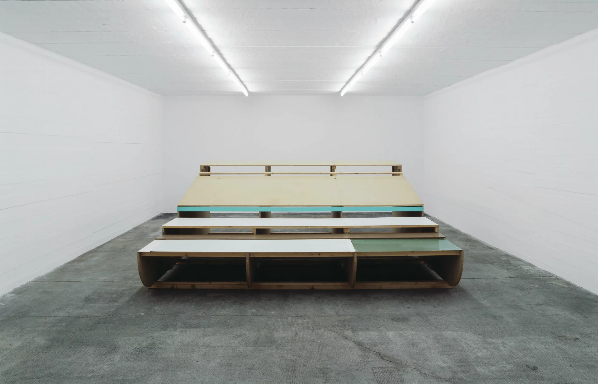 Rauminstallation aus Holzplatten und bunten Flächen, gestapelt zu einer tribünenartigen Struktur in weißem Ausstellungsraum.