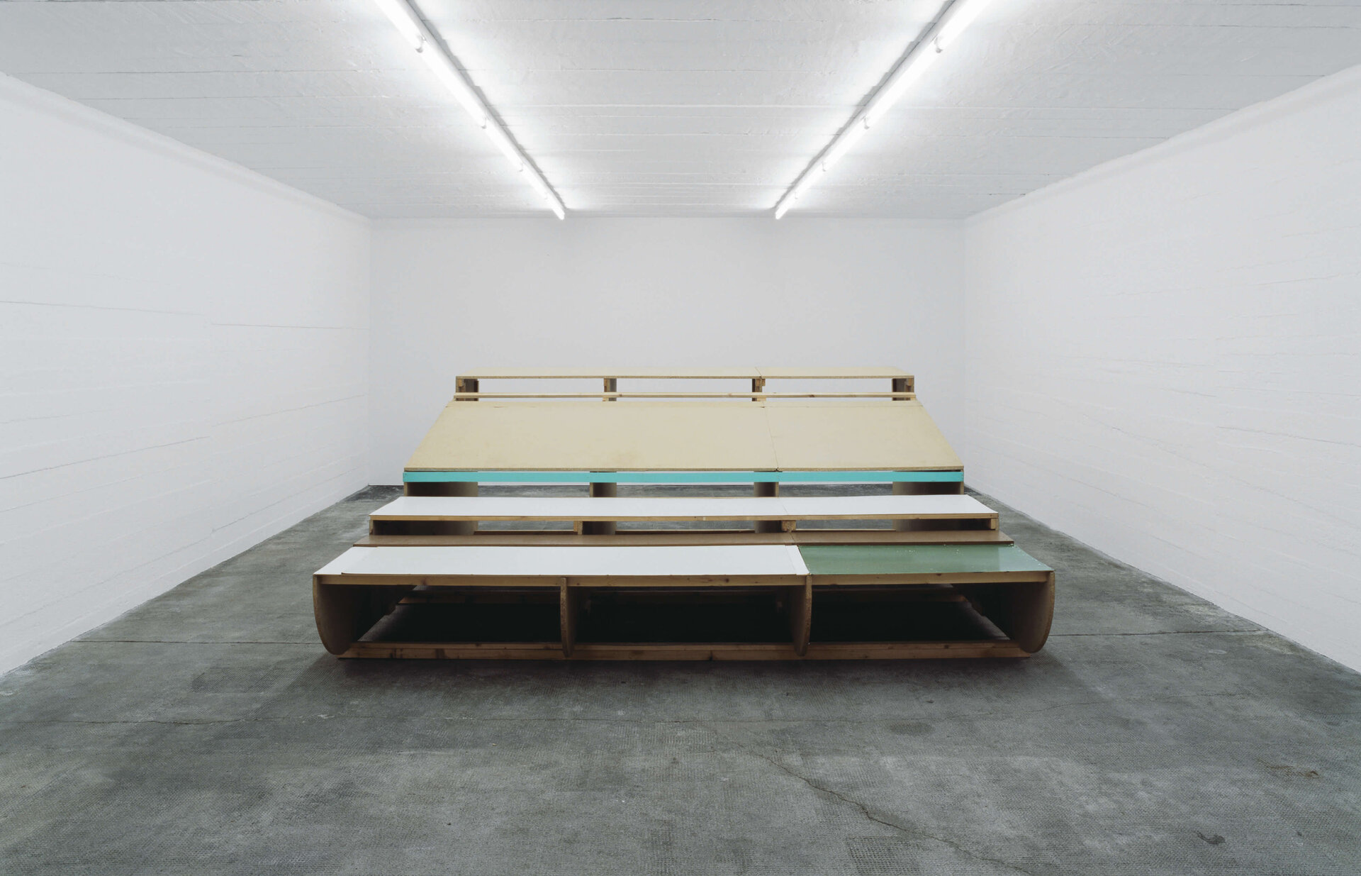 Rauminstallation aus Holzplatten und bunten Flächen, gestapelt zu einer tribünenartigen Struktur in weißem Ausstellungsraum.