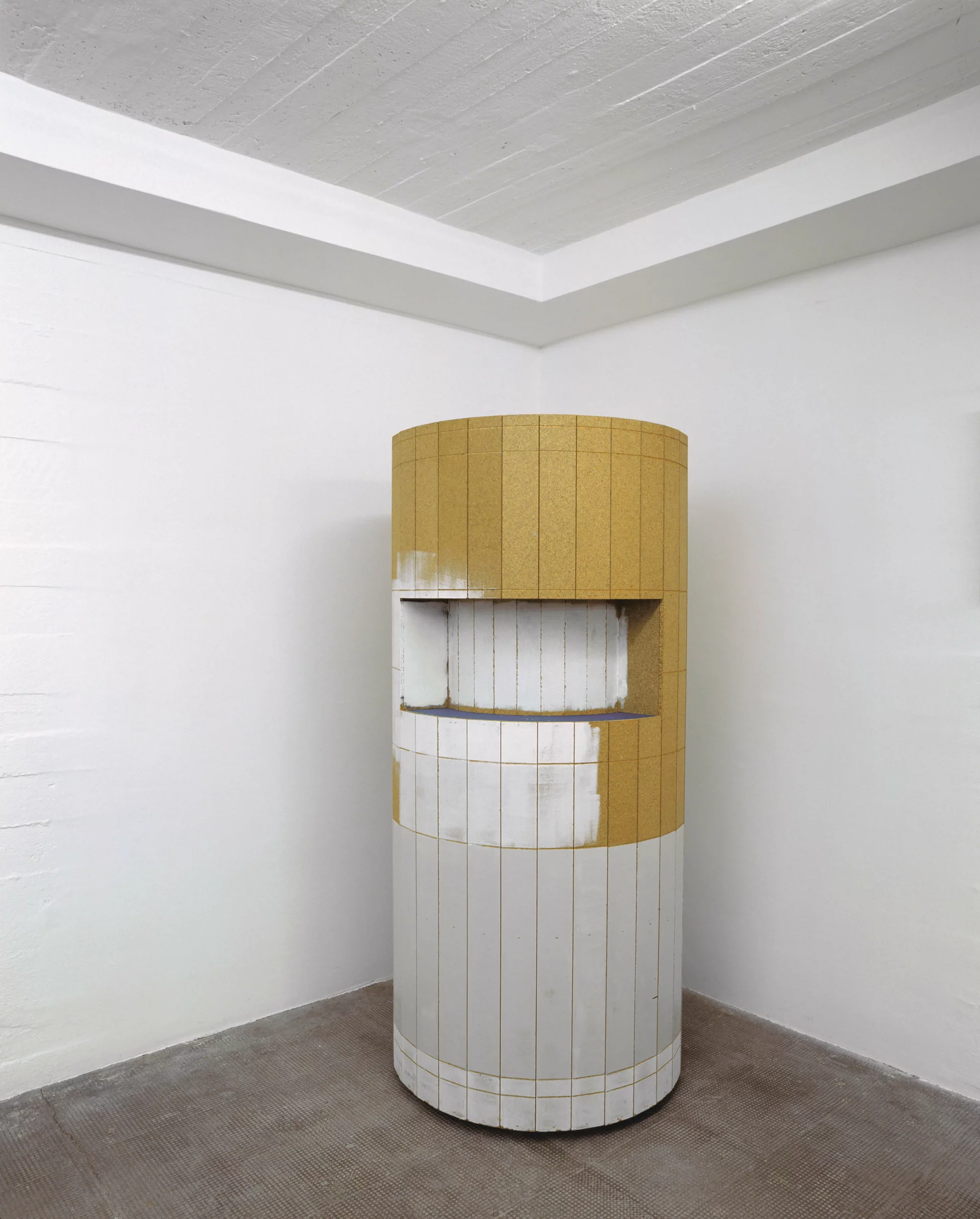 Zylindrische Skulptur mit gelben und weißen Kachelflächen, mit eingelassener Öffnung in einem weißen Ausstellungsraum.