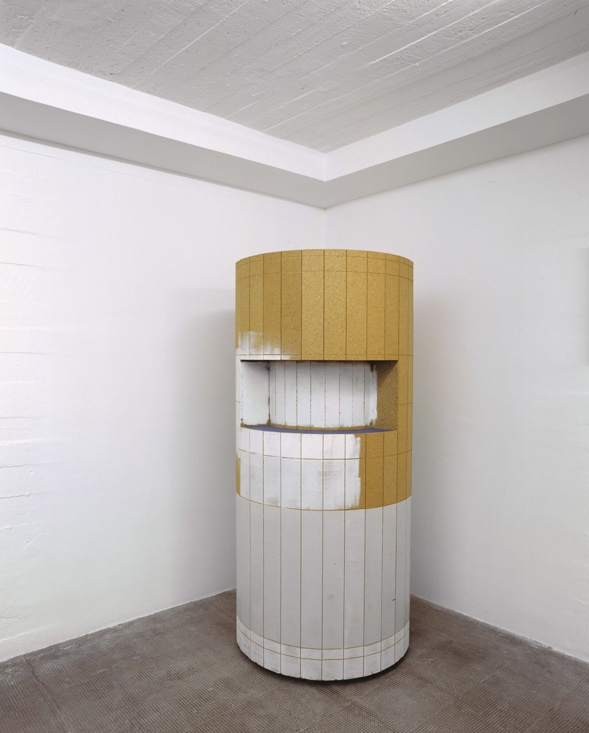 Zylindrische Skulptur mit gelben und weißen Kachelflächen, mit eingelassener Öffnung in einem weißen Ausstellungsraum.