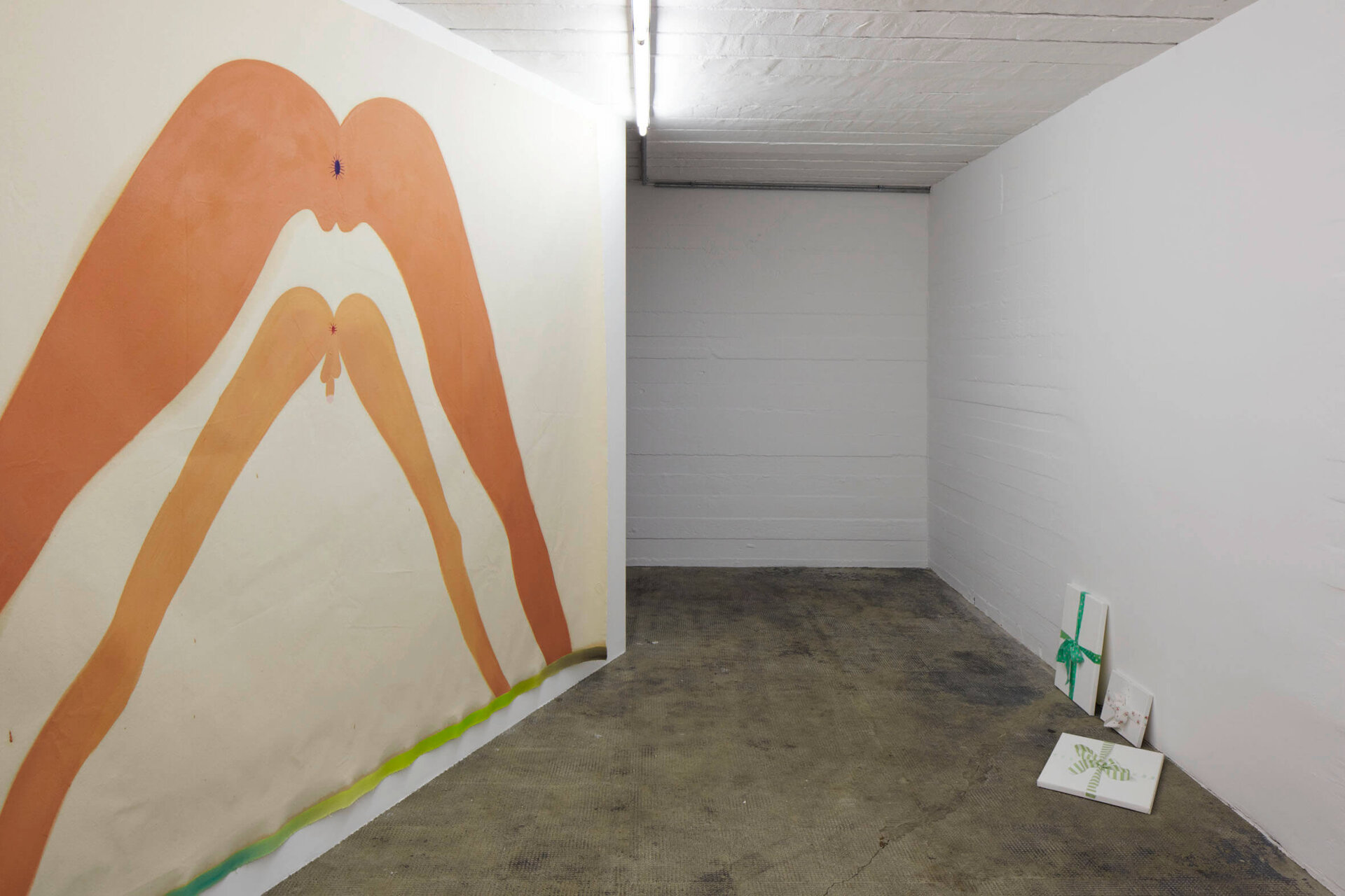 Großflächige Malerei von zwei übereinandergelegten, abstrahierten Körperpaaren an der Wand; auf dem Boden im rechten Bildbereich stehen und liegen kleine, weiß grundierte Leinwände mit grünen Schleifen.