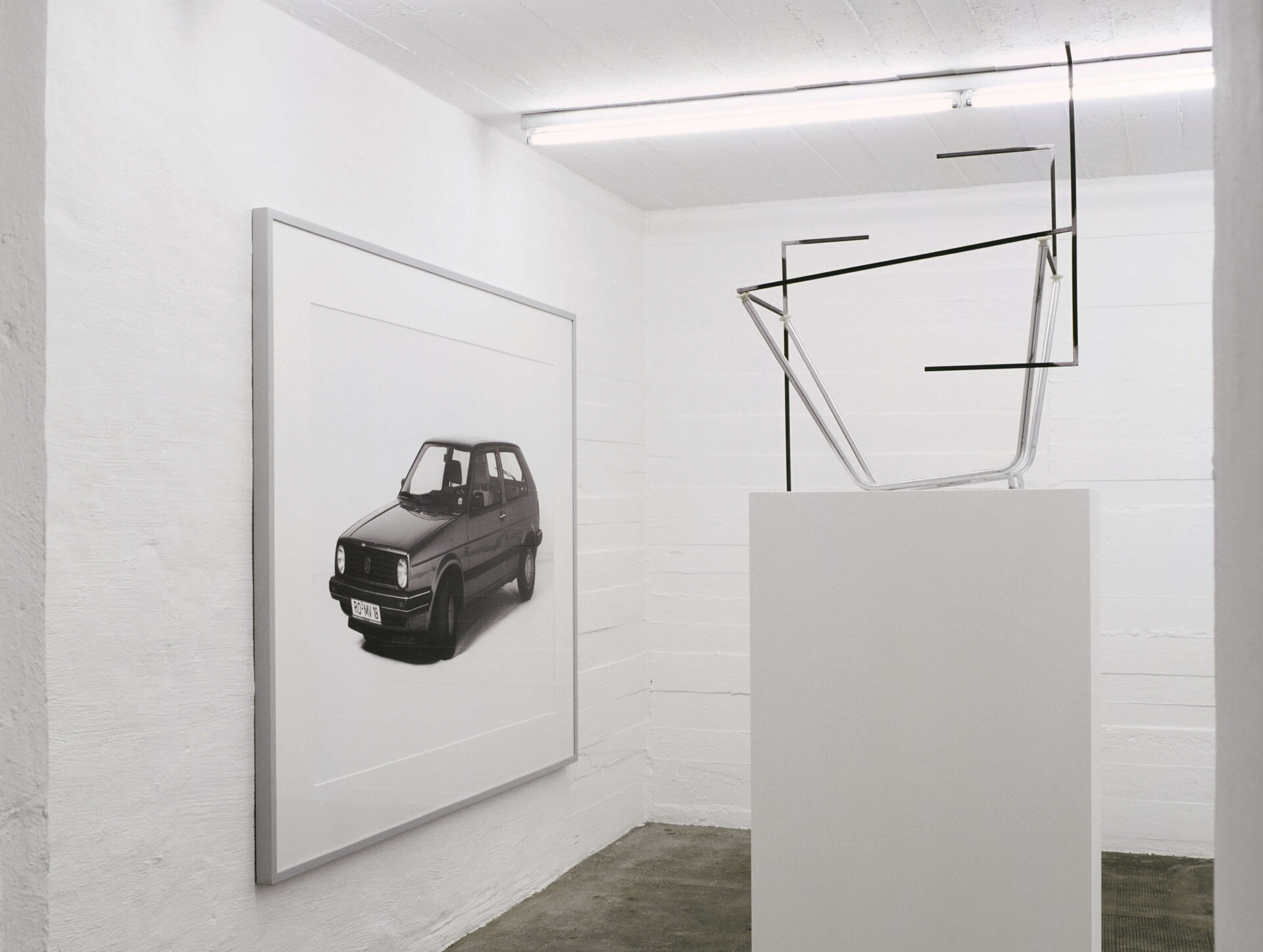 Foto eines schwarzen VW Golf neben einer minimalistischen Skulptur aus Fahrradteilen in einem weißen Ausstellungsraum.
