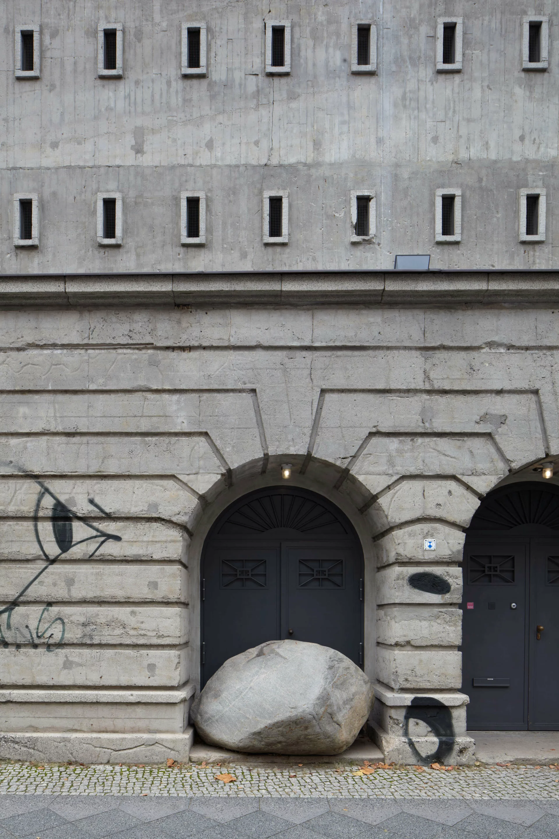 Großer Naturstein liegt scheinbar zufällig vor dem mittleren Torbogen des Boros Bunkers in Berlin; der Stein blockiert einen der Eingänge, während kleine architektonische Details und Graffiti die rohe Umgebung betonen.