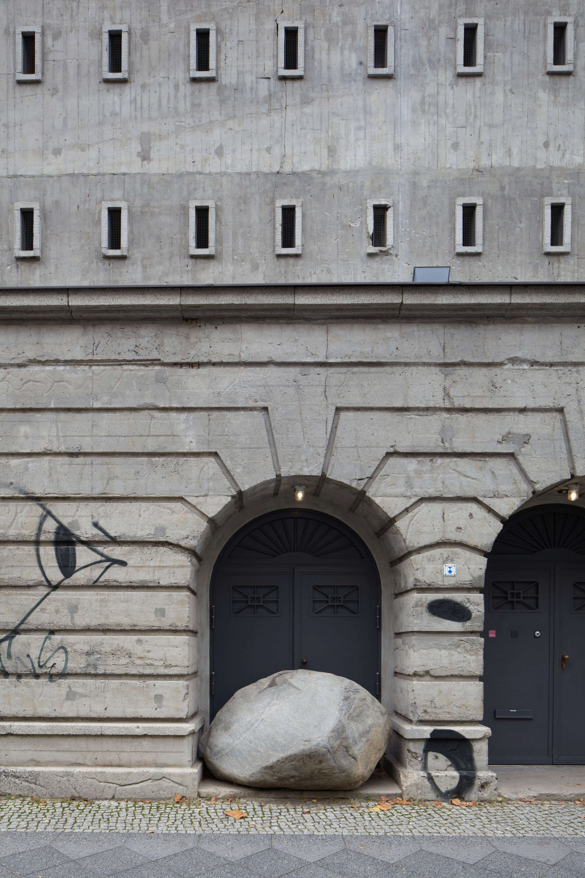 Großer Naturstein liegt scheinbar zufällig vor dem mittleren Torbogen des Boros Bunkers in Berlin; der Stein blockiert einen der Eingänge, während kleine architektonische Details und Graffiti die rohe Umgebung betonen.