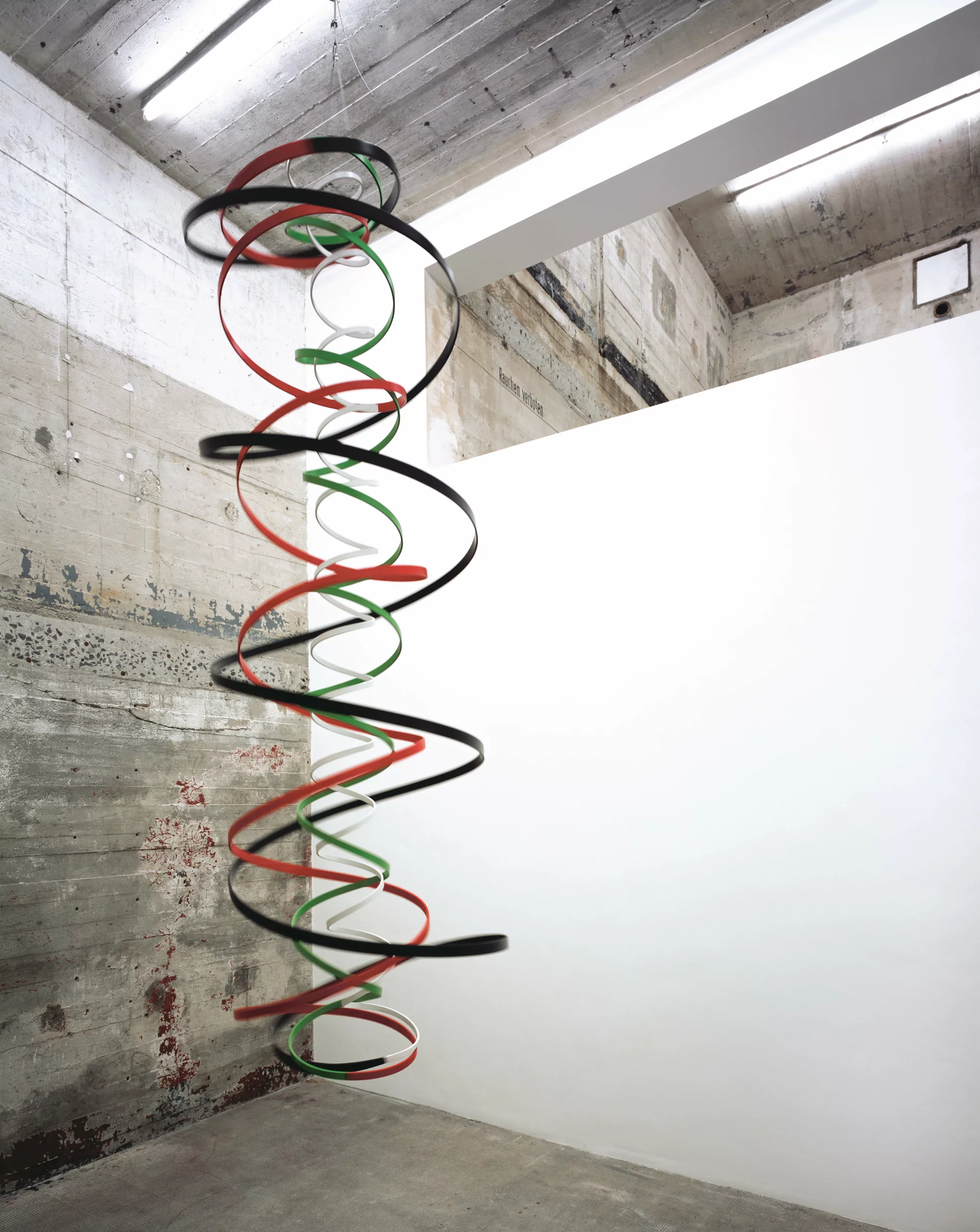 Hängende Spiralskulptur aus farbigen Bändern (rot, grün, schwarz, weiß) in einem Ausstellungsraum mit Betonwand.