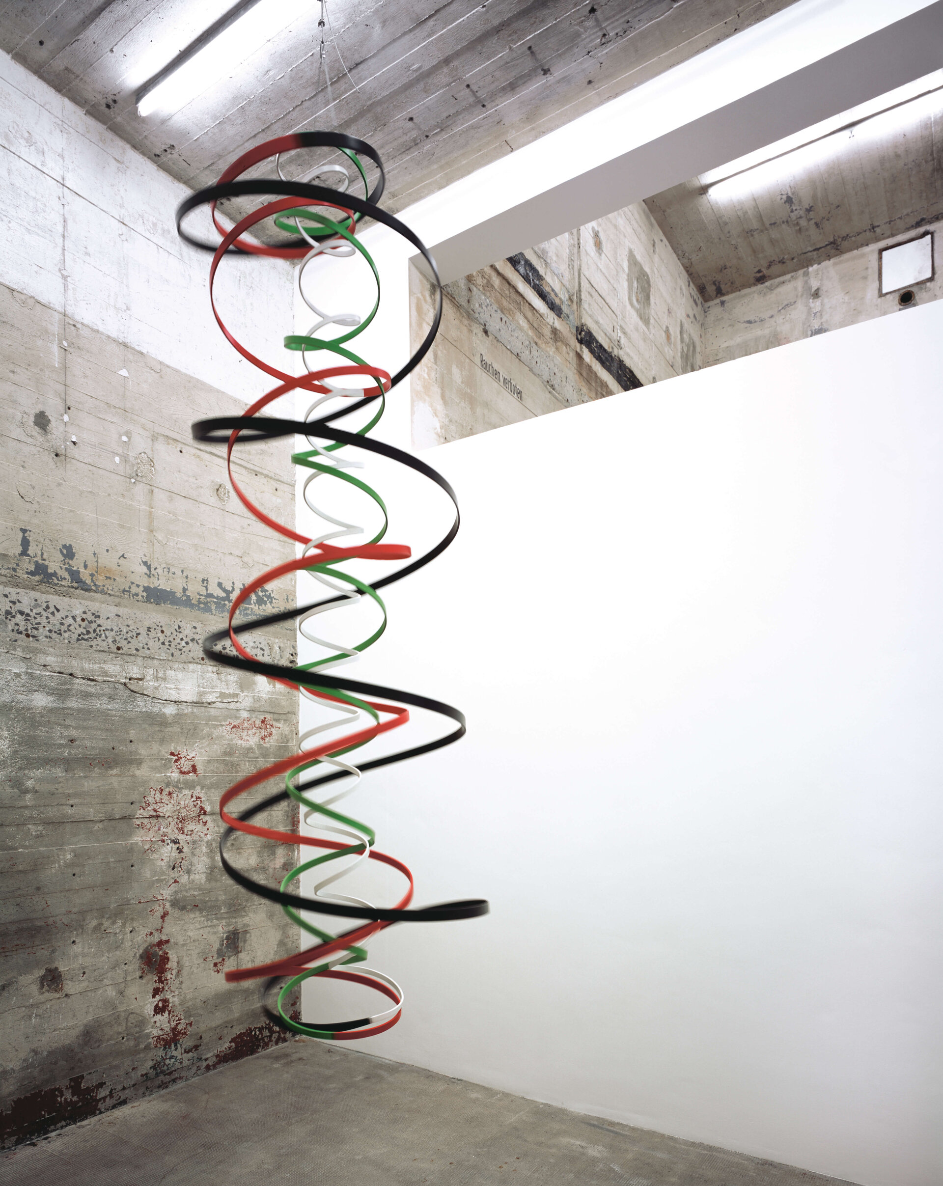 Hängende Spiralskulptur aus farbigen Bändern (rot, grün, schwarz, weiß) in einem Ausstellungsraum mit Betonwand.