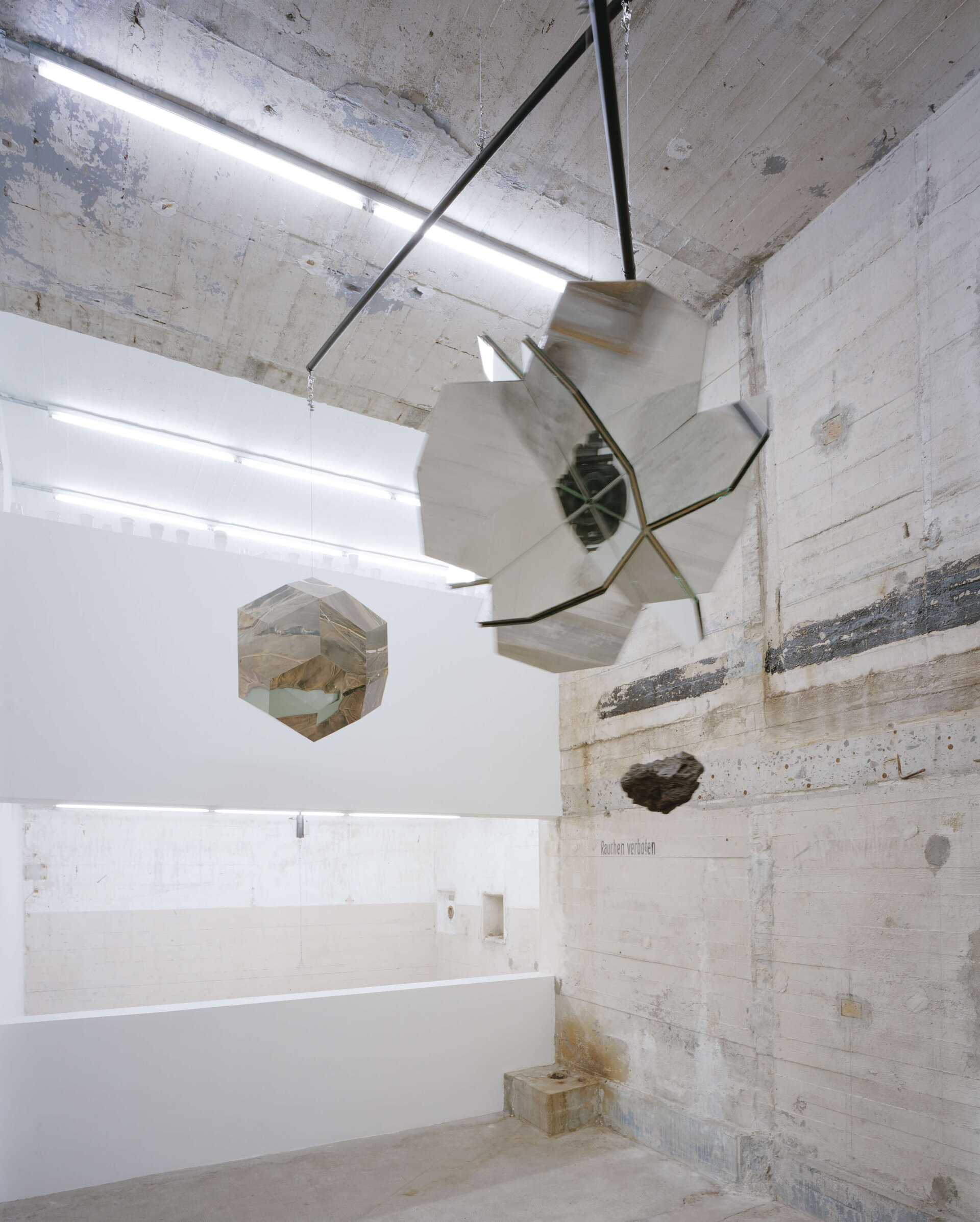 Spiegelnde geometrische Skulpturen und ein schwebender Stein in einem roh belassenen Ausstellungsraum mit Betonwänden.