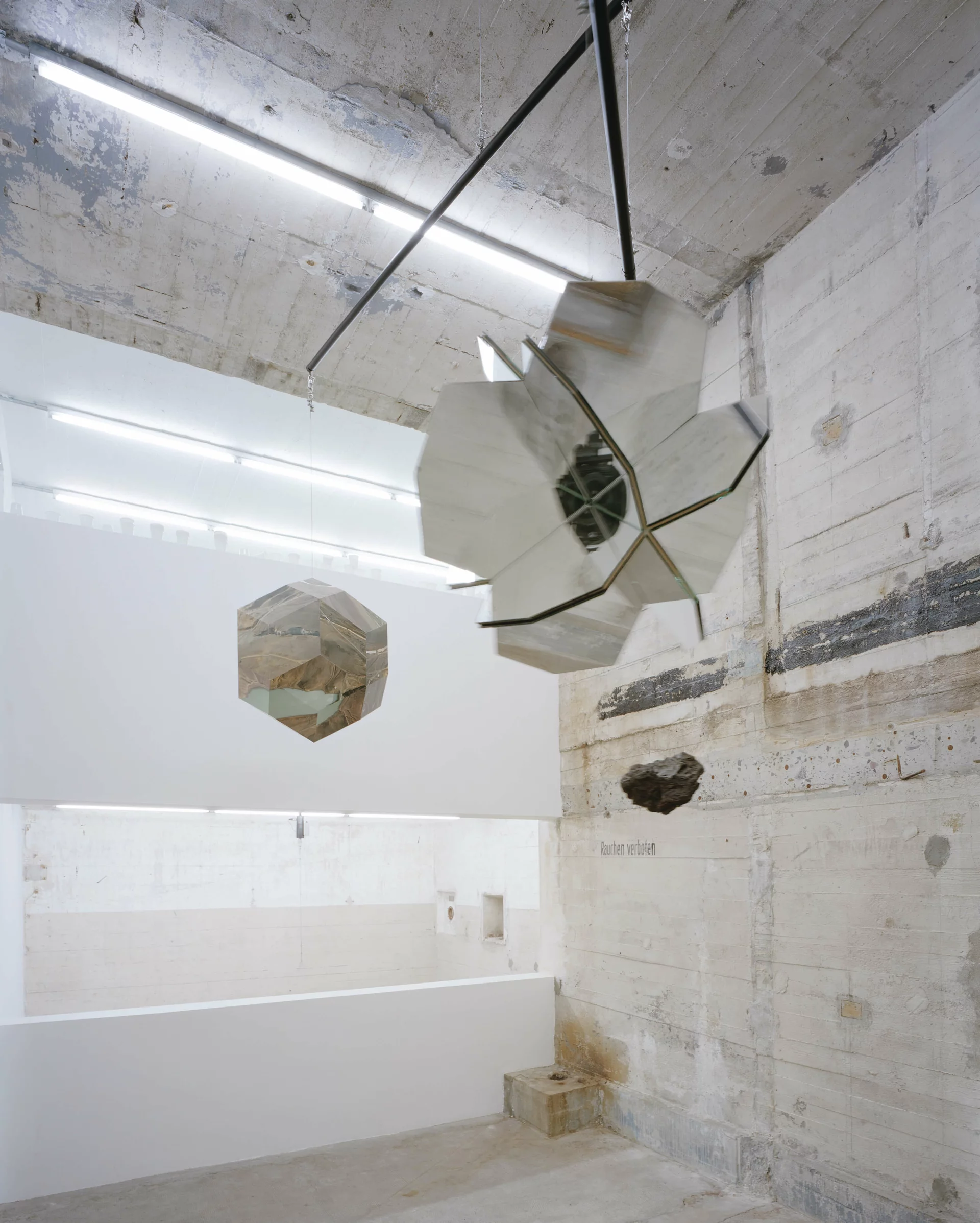 Spiegelnde geometrische Skulpturen und ein schwebender Stein in einem roh belassenen Ausstellungsraum mit Betonwänden.