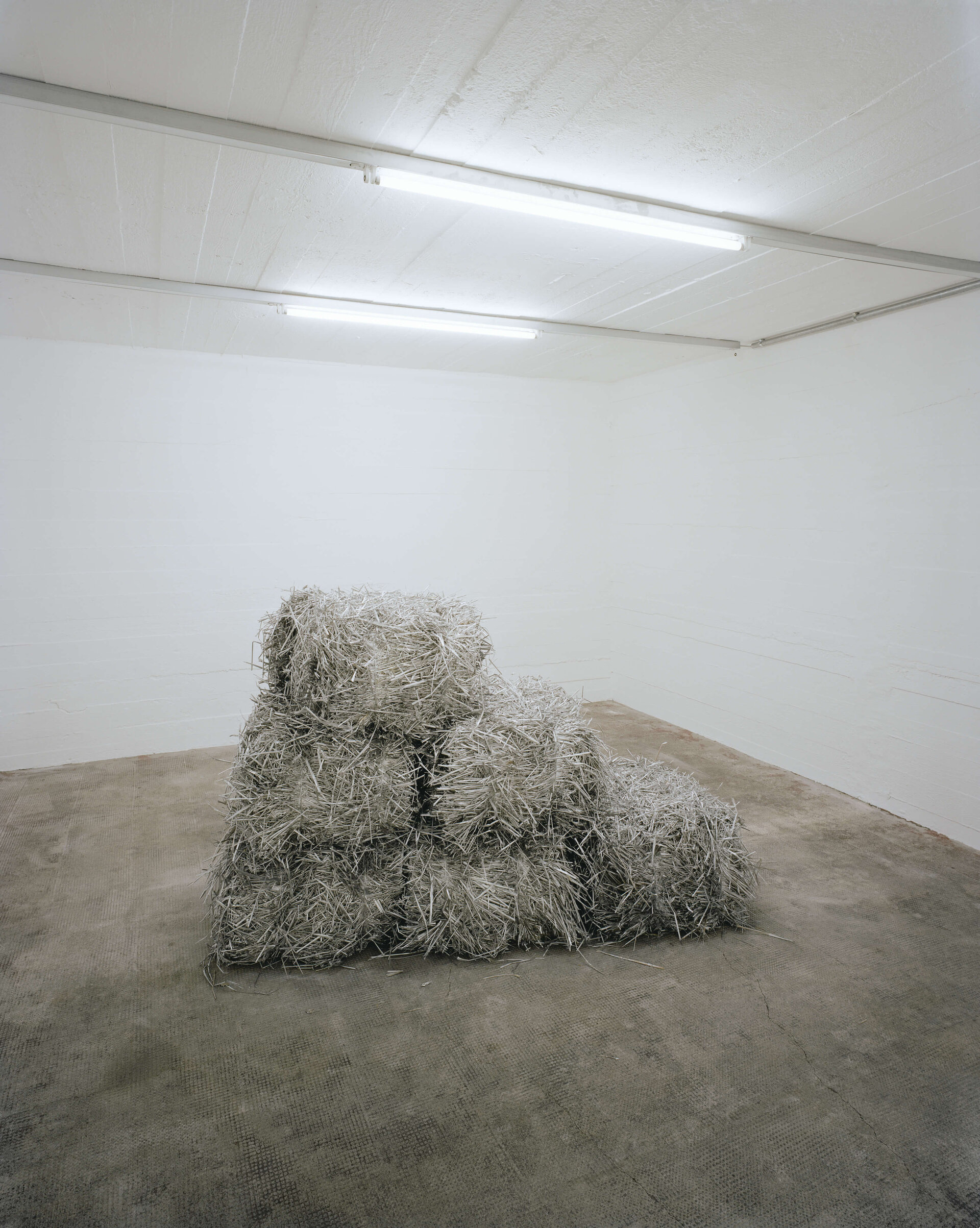 Pyramidenförmig gestapelte, silberglänzende Strohballen in einem weißen, kahlen Raum mit Betonboden.