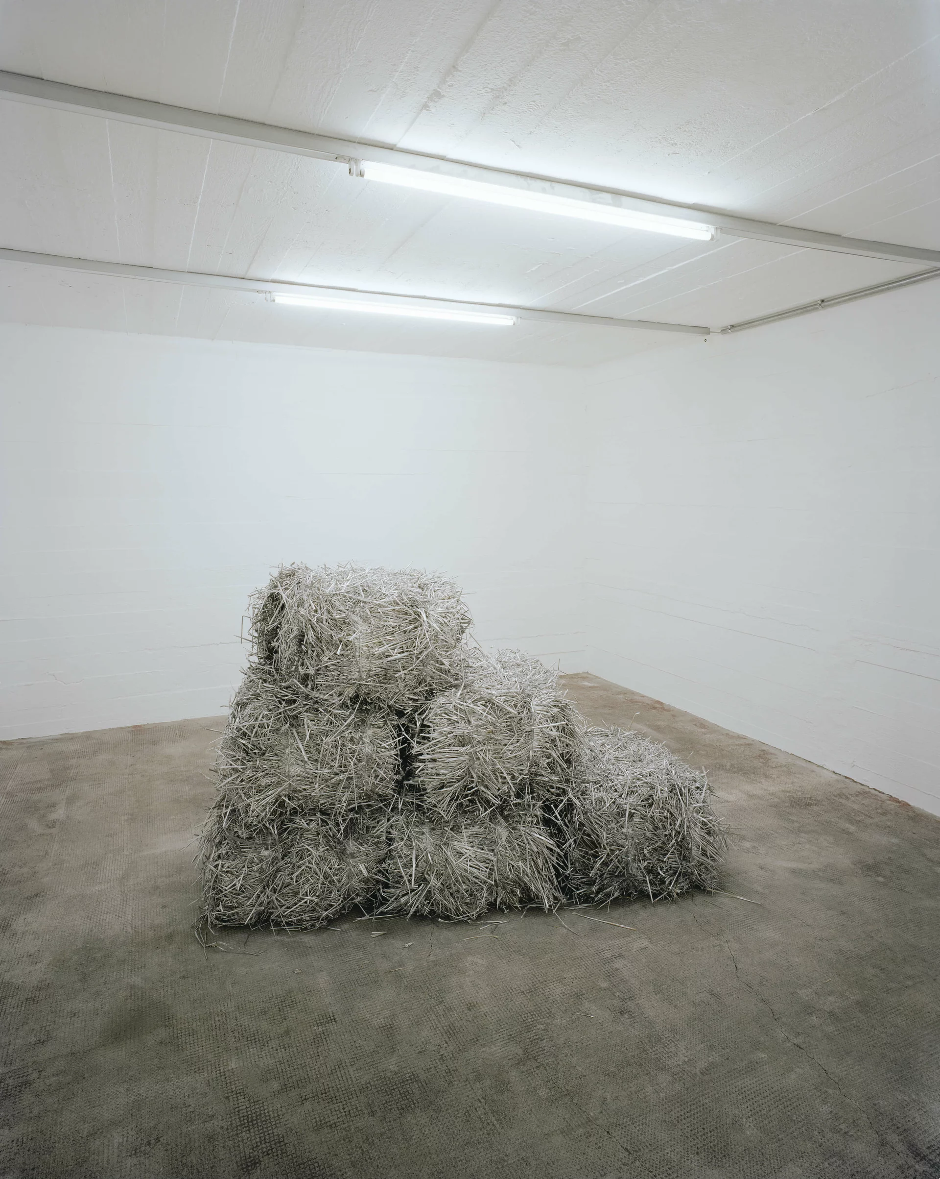 Pyramidenförmig gestapelte, silberglänzende Strohballen in einem weißen, kahlen Raum mit Betonboden.