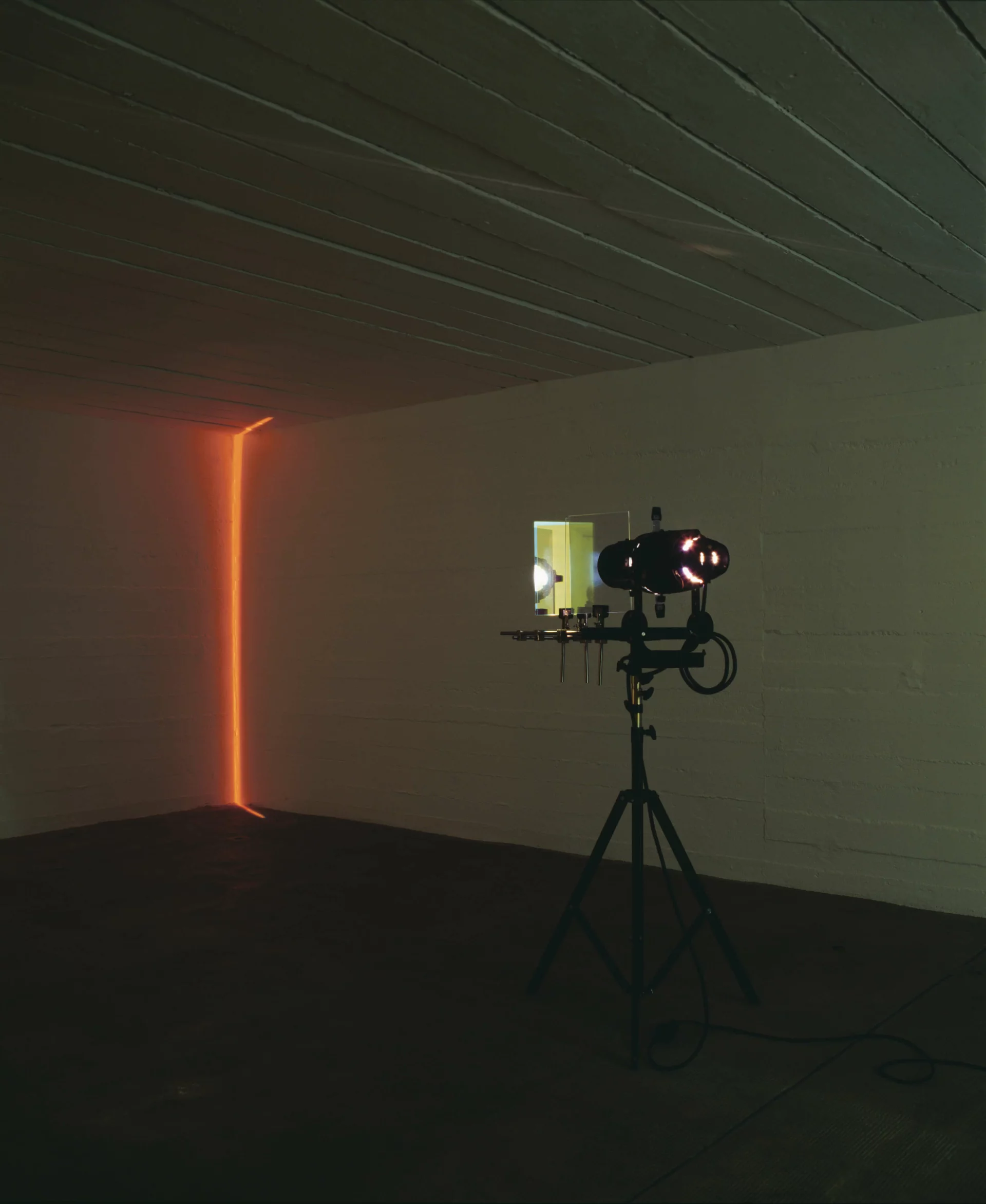 Lichtprojektion in dunklem Raum, bei der ein schmaler, vertikaler Lichtstrahl eine Ecke in warmem Orange illuminiert.