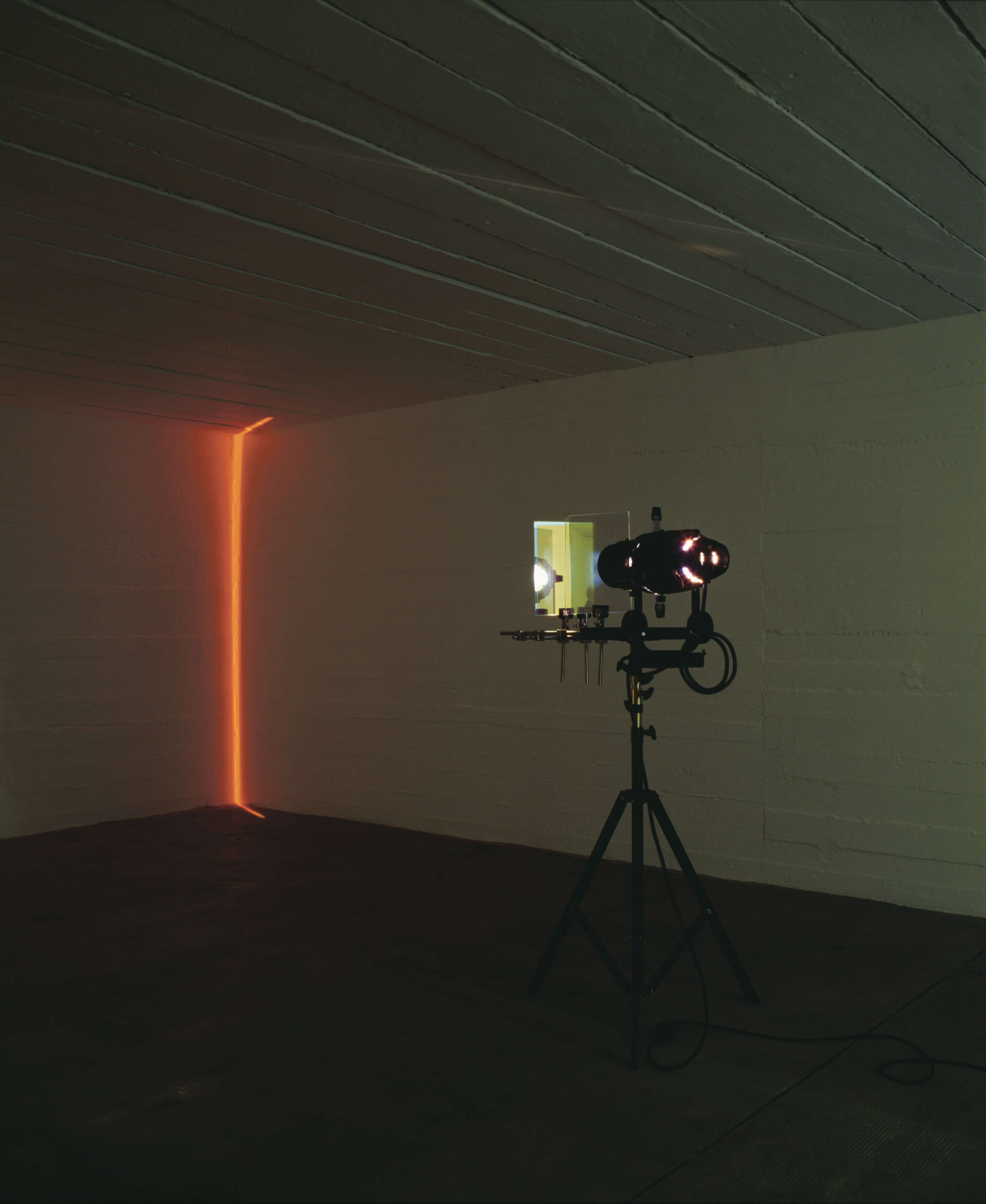 Lichtprojektion in dunklem Raum, bei der ein schmaler, vertikaler Lichtstrahl eine Ecke in warmem Orange illuminiert.