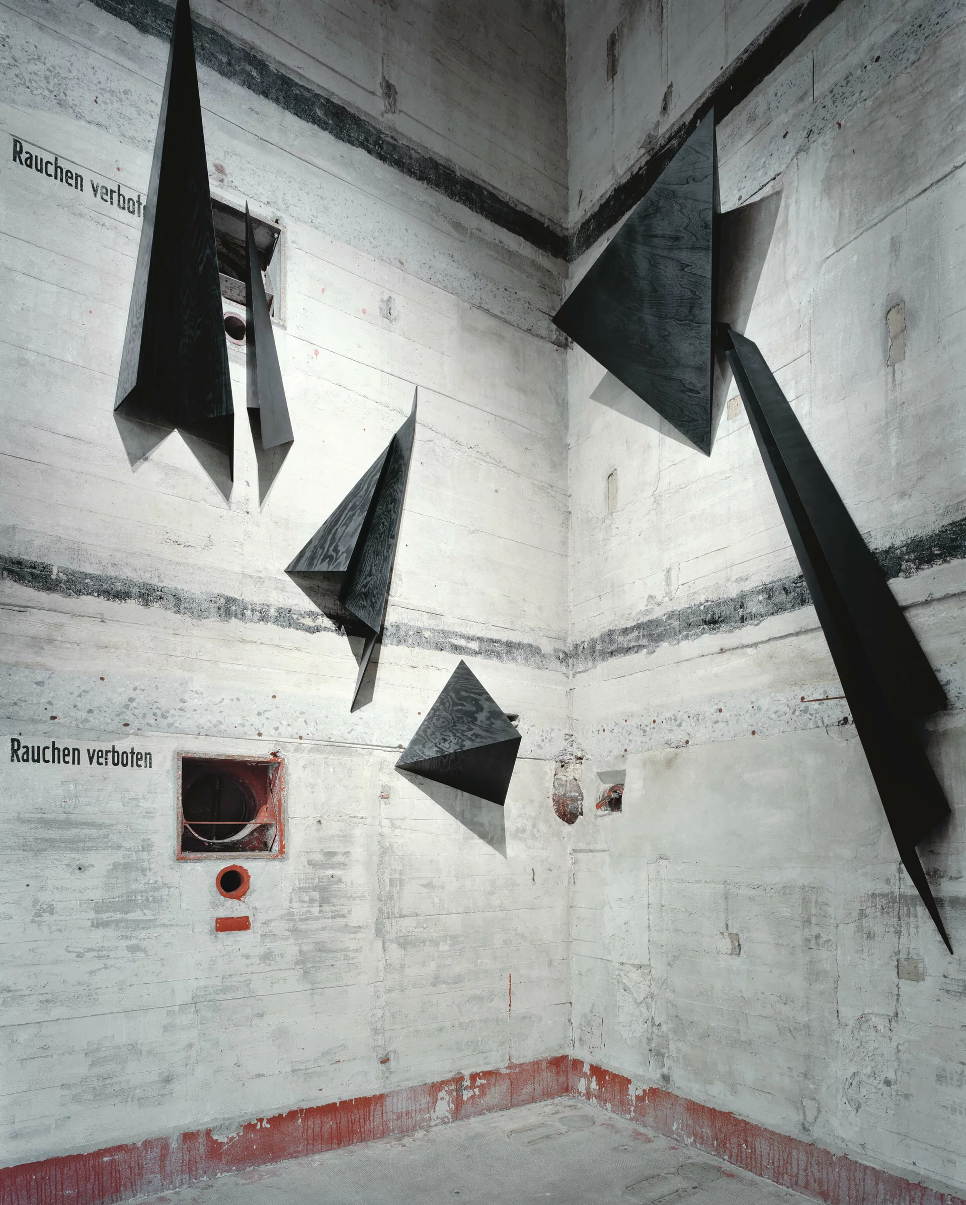 Schwarze, gefaltete Metallskulpturen hängen in einem hohen, rauen Betonraum mit der Aufschrift „Rauchen verboten“.