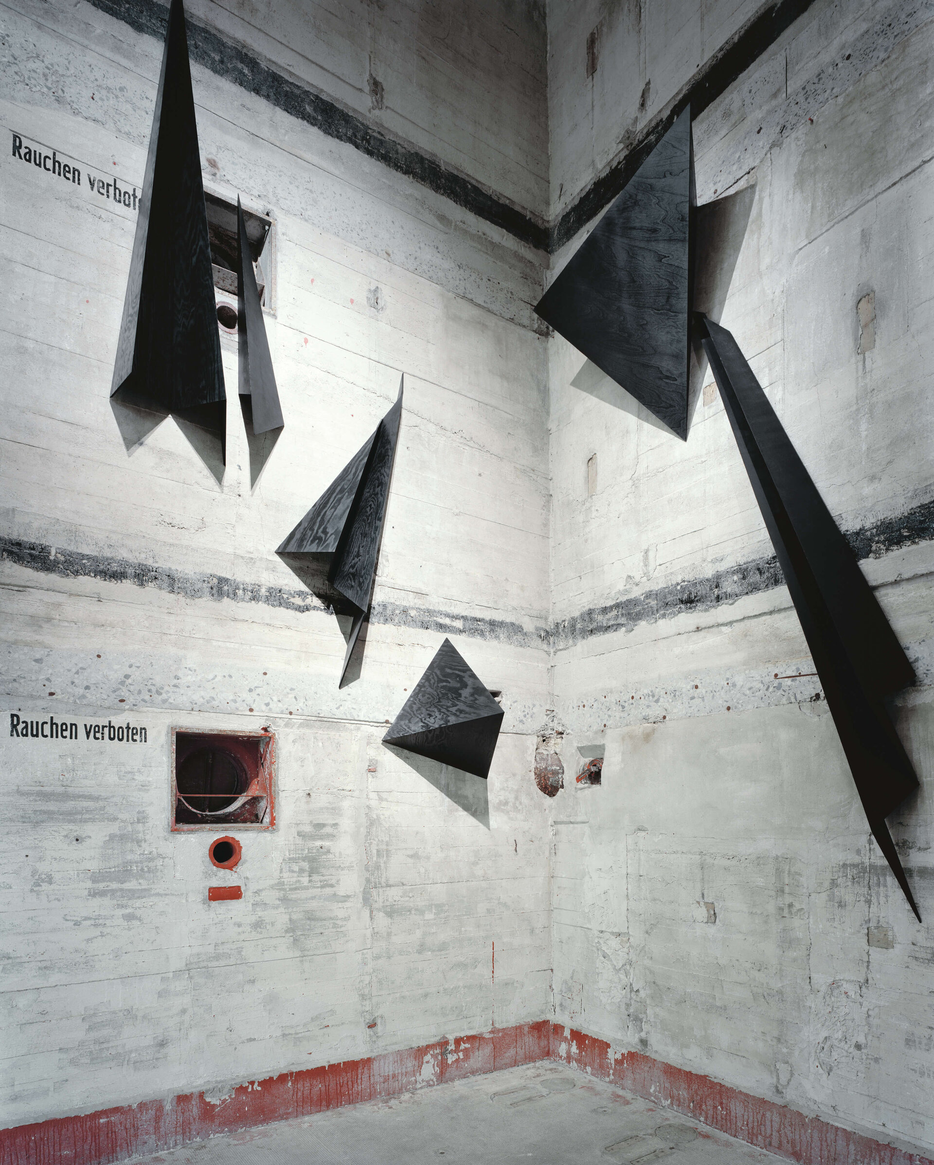 Schwarze, gefaltete Metallskulpturen hängen in einem hohen, rauen Betonraum mit der Aufschrift „Rauchen verboten“.