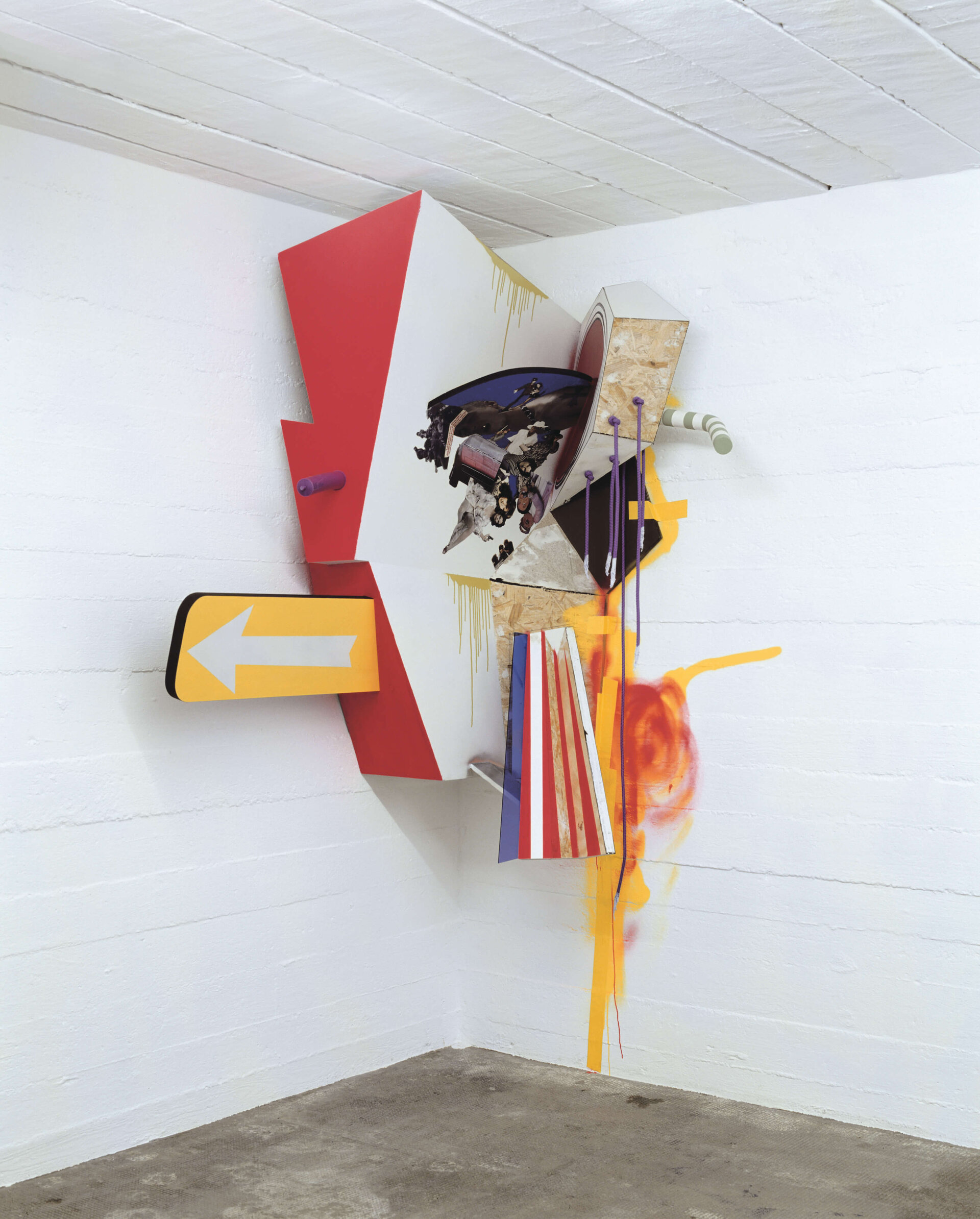 Bunte, kantige Wandinstallation mit Collage-Elementen, Schildern und Farbspritzern in einer weißen Raumecke.