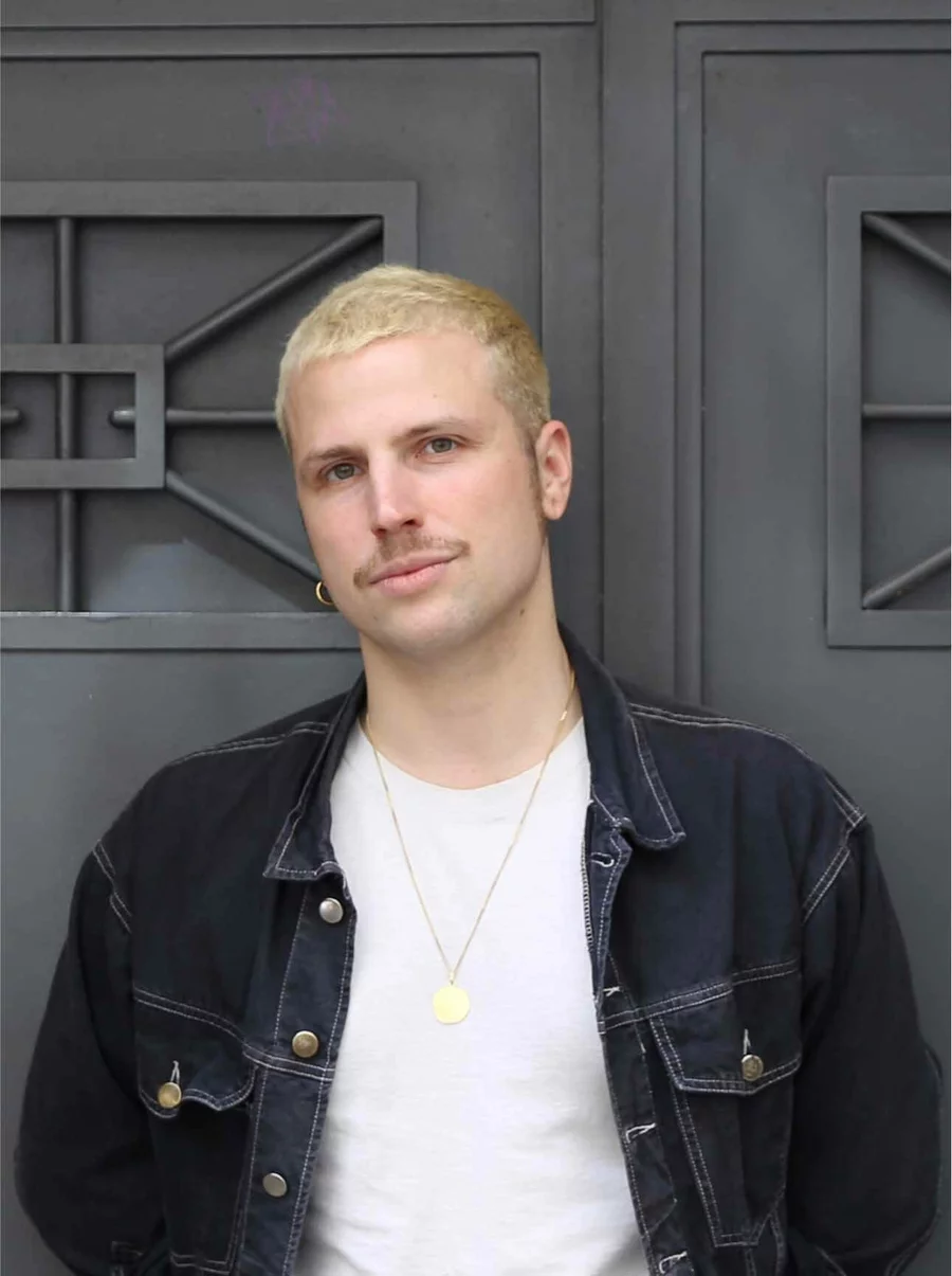 Porträt von Justus mit blondem Kurzhaarschnitt, weißem Shirt und Jeansjacke vor grauer Metalltür.