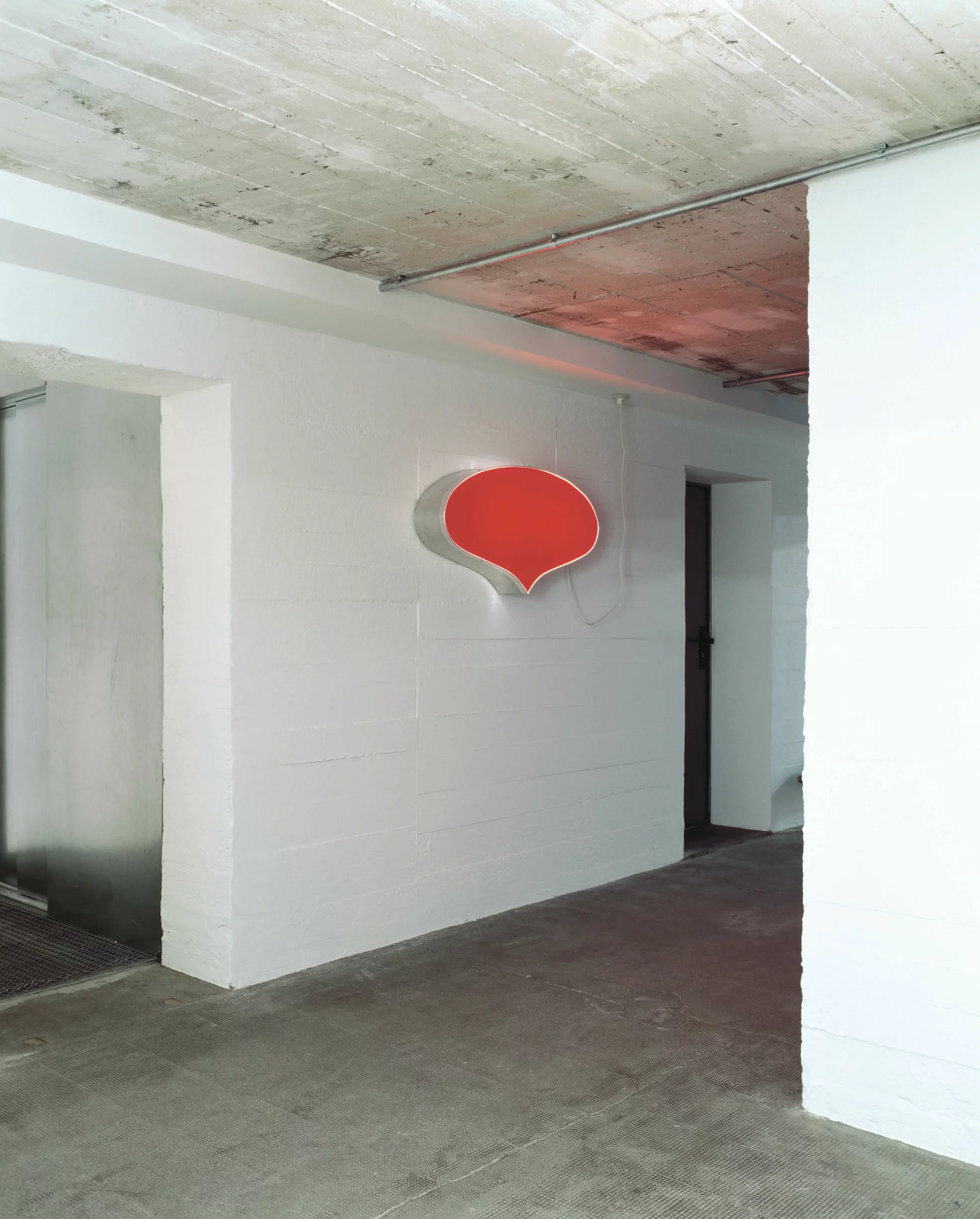 Rotes Leuchtobjekt in Form einer Sprechblase an weißer Wand in industrieller Umgebung mit Betonboden und Decke.