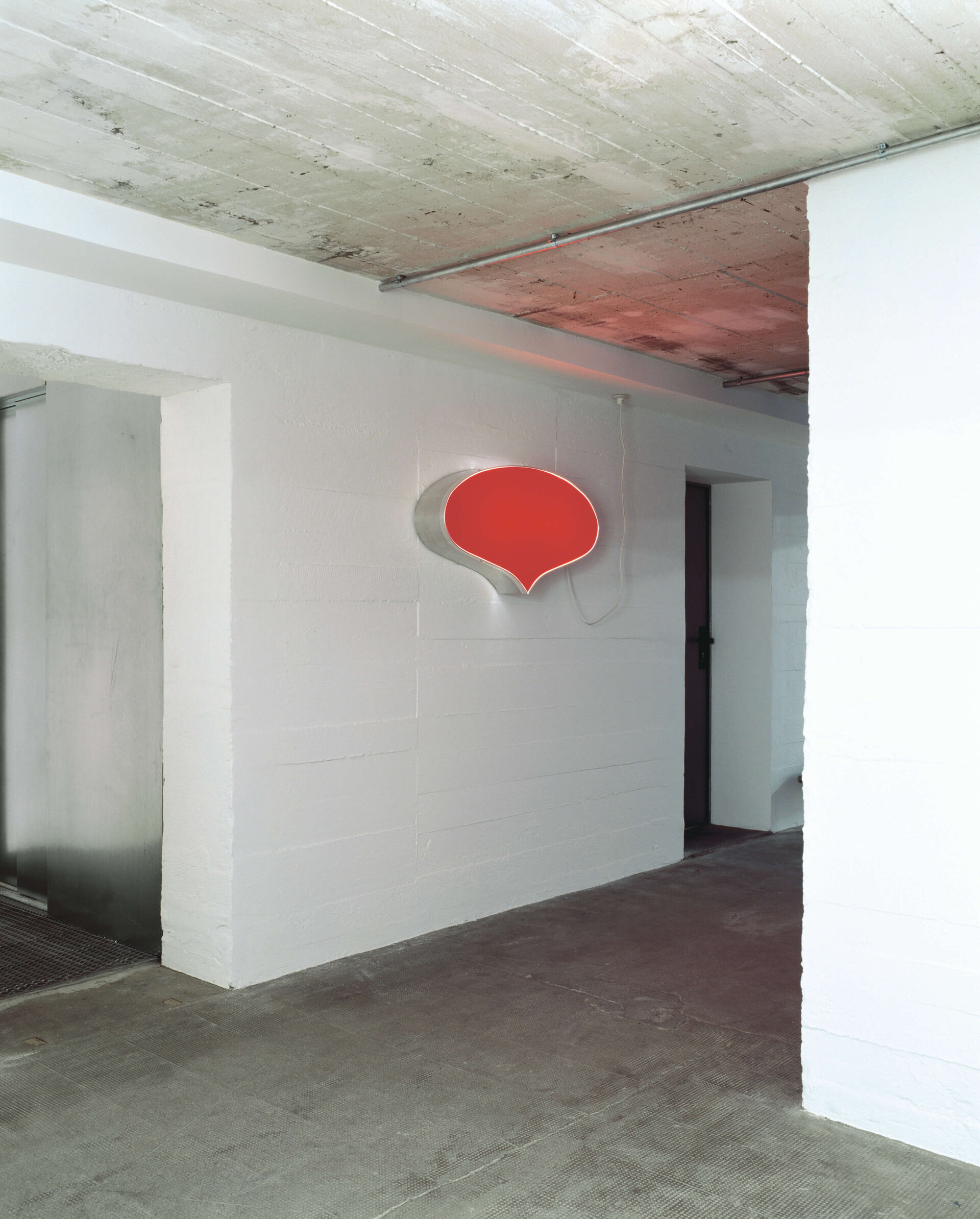 Rotes Leuchtobjekt in Form einer Sprechblase an weißer Wand in industrieller Umgebung mit Betonboden und Decke.