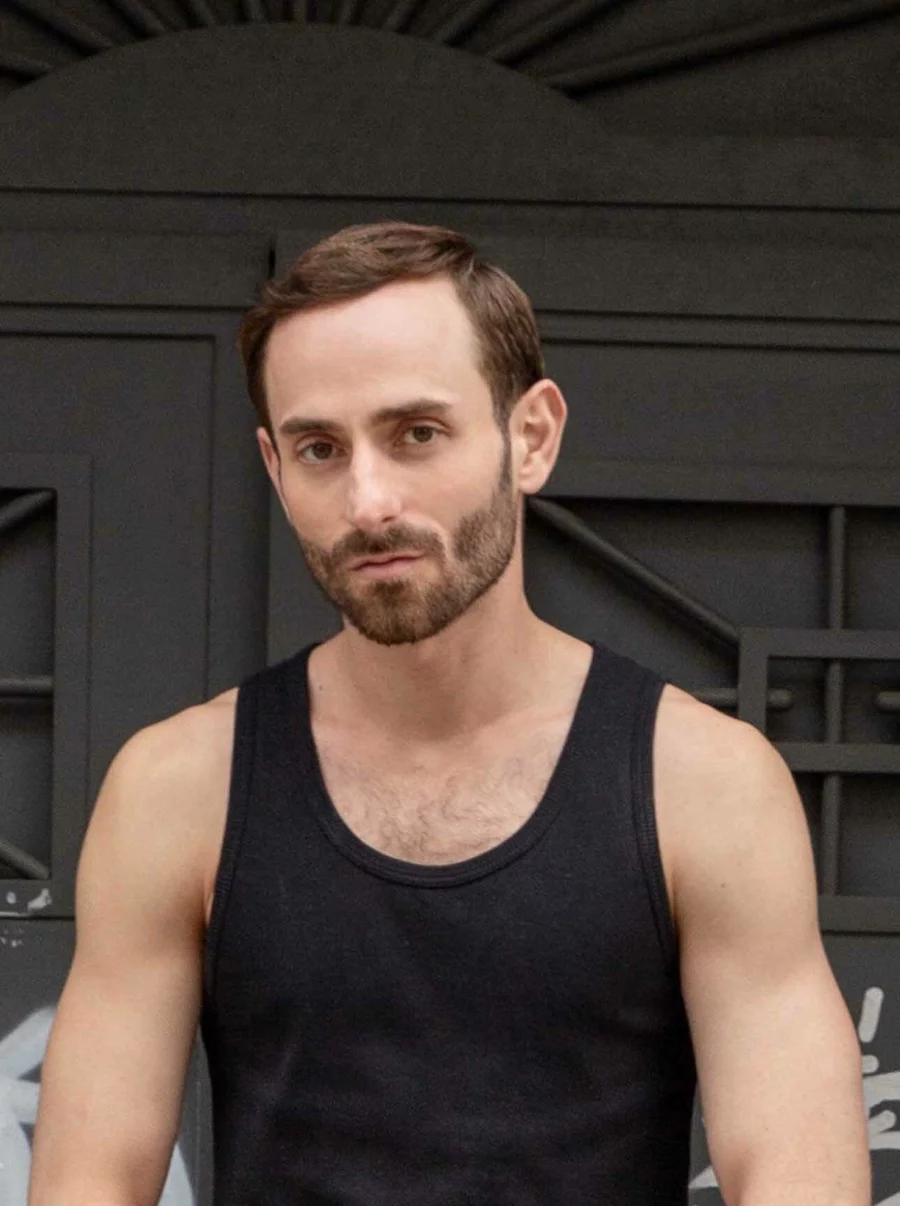 Jean-Christian im schwarzen Tanktop vor strukturierter Tür mit geometrischen Details, direkter Blick in die Kamera.