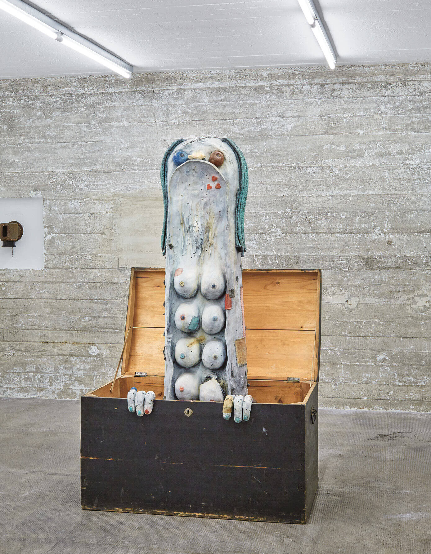 Bunte Skulptur mit vielen Brüsten und menschlichem Gesicht ragt aus einer Holztruhe in einem rauen Ausstellungsraum empor.