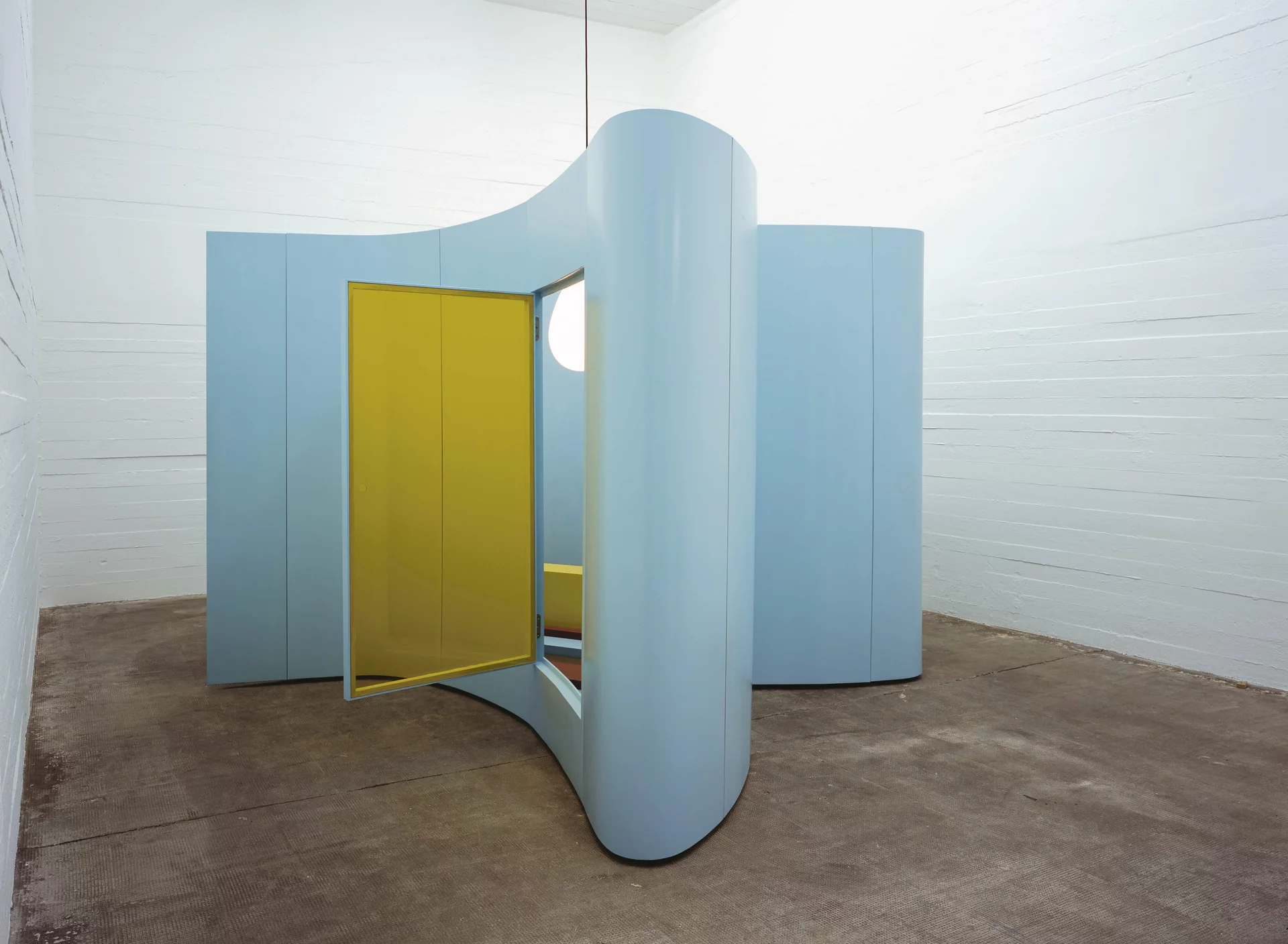 Geschwungene, pastellblaue Raumskulptur mit geöffnetem gelben Türelement in weißem Ausstellungsraum.
