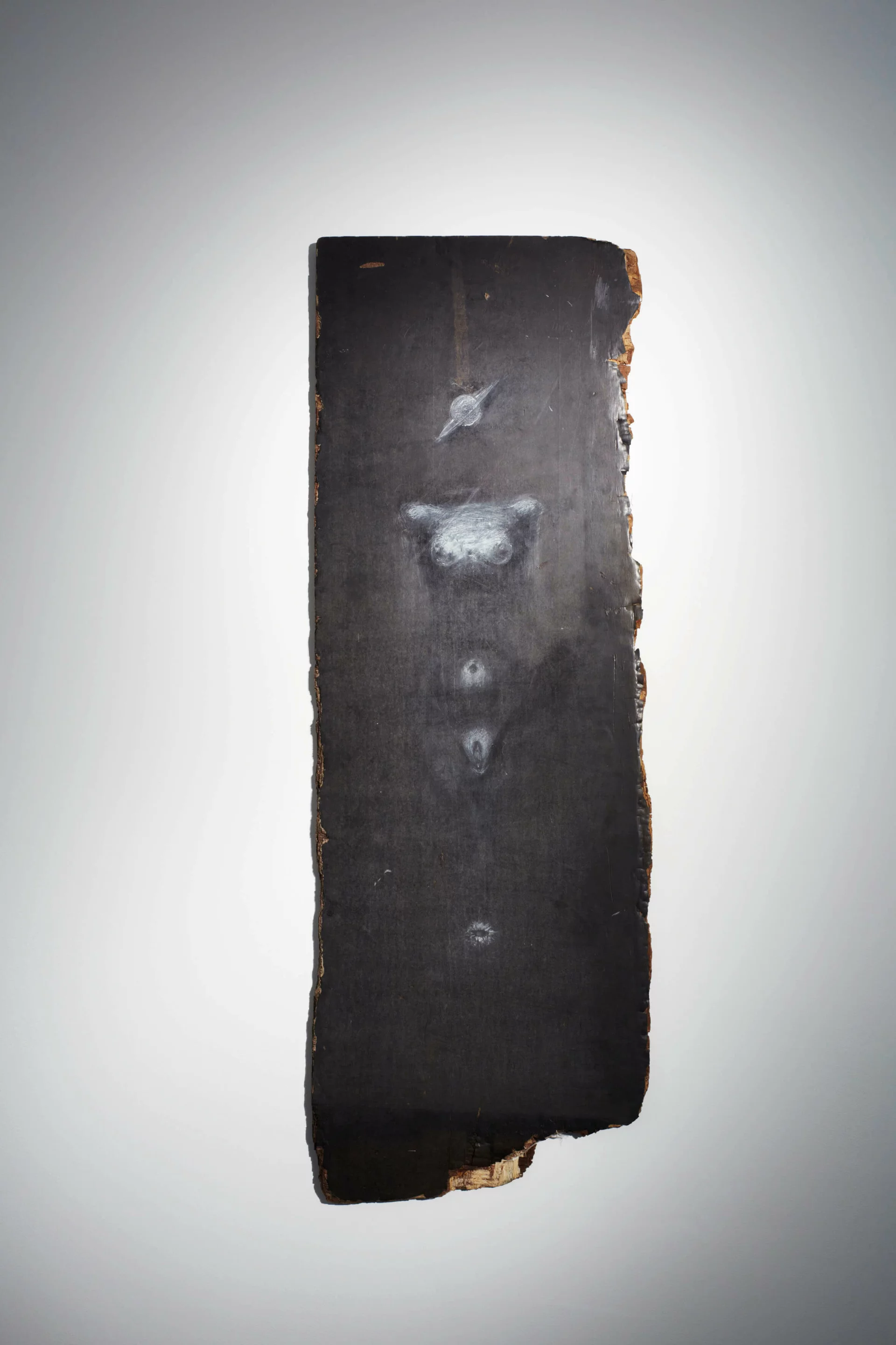 Vertikal gehängtes, dunkles Holzbrett mit weißer, schemenhafter Zeichnung eines menschlichen Torsos und angedeuteten Symbolen; organisch unregelmäßiger Rand.