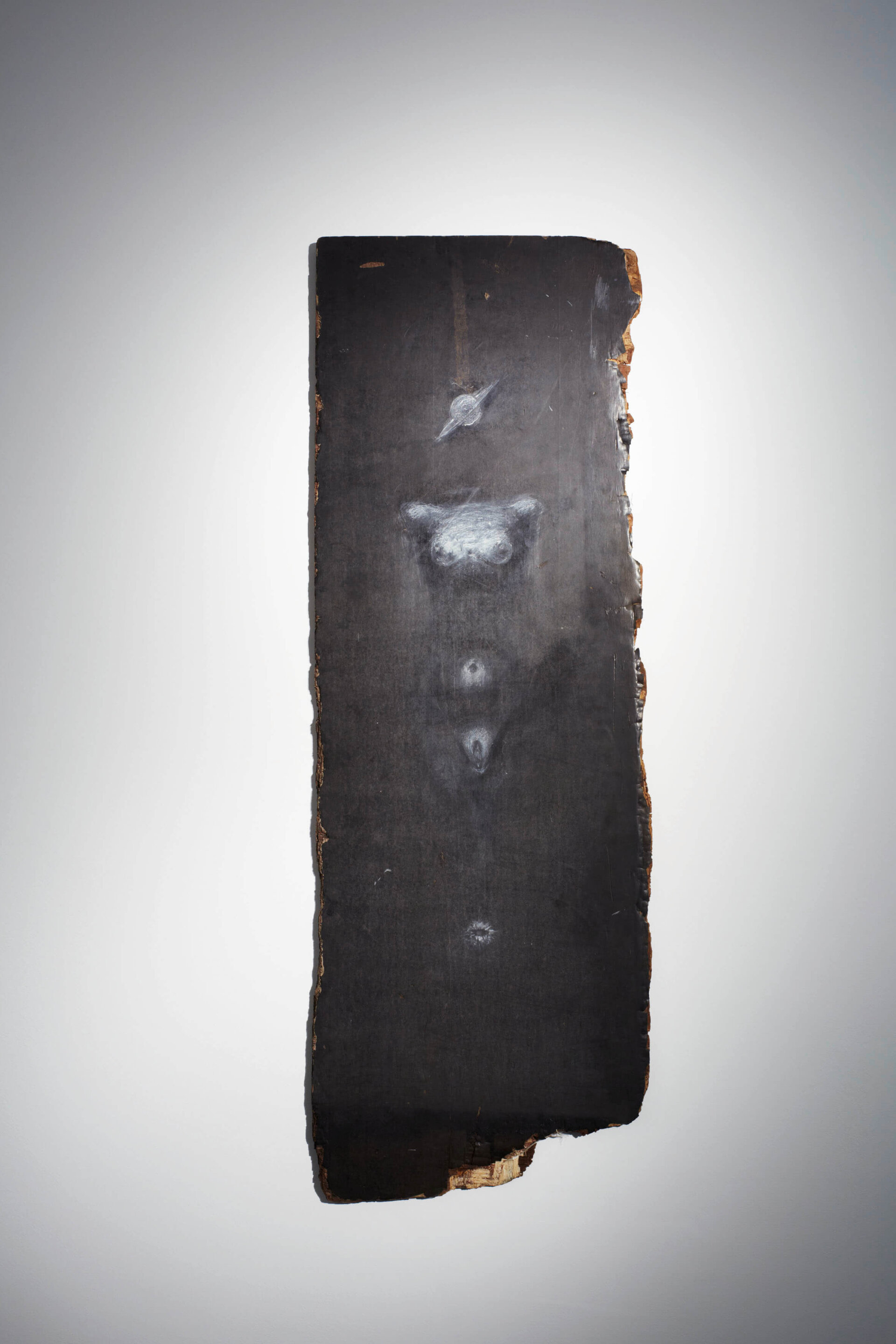 Vertikal gehängtes, dunkles Holzbrett mit weißer, schemenhafter Zeichnung eines menschlichen Torsos und angedeuteten Symbolen; organisch unregelmäßiger Rand.