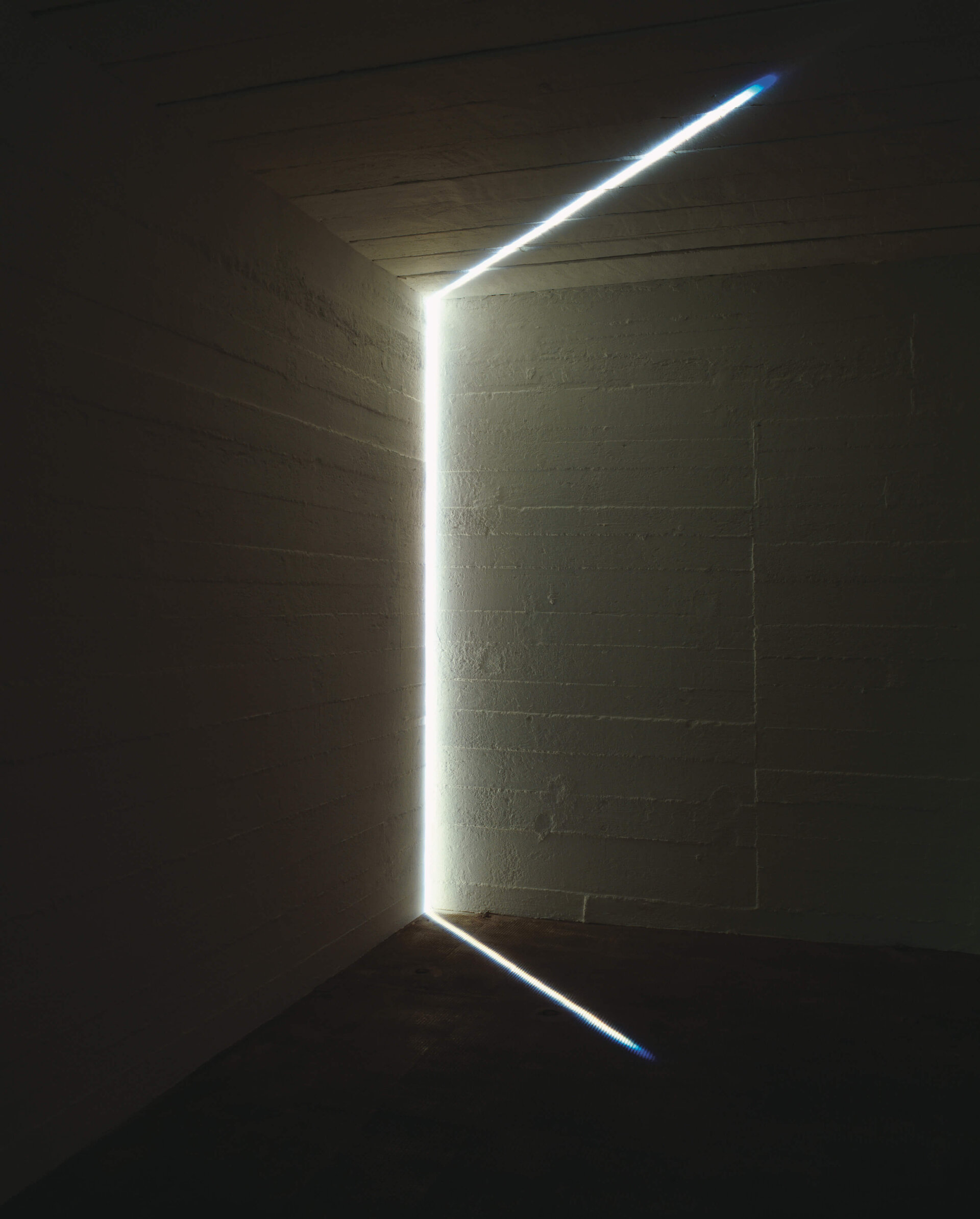 Feiner weißer Lichtstrahl durchzieht die Ecke eines dunklen Raumes und wirft geometrische Lichtlinien auf Wände, Boden und Decke.