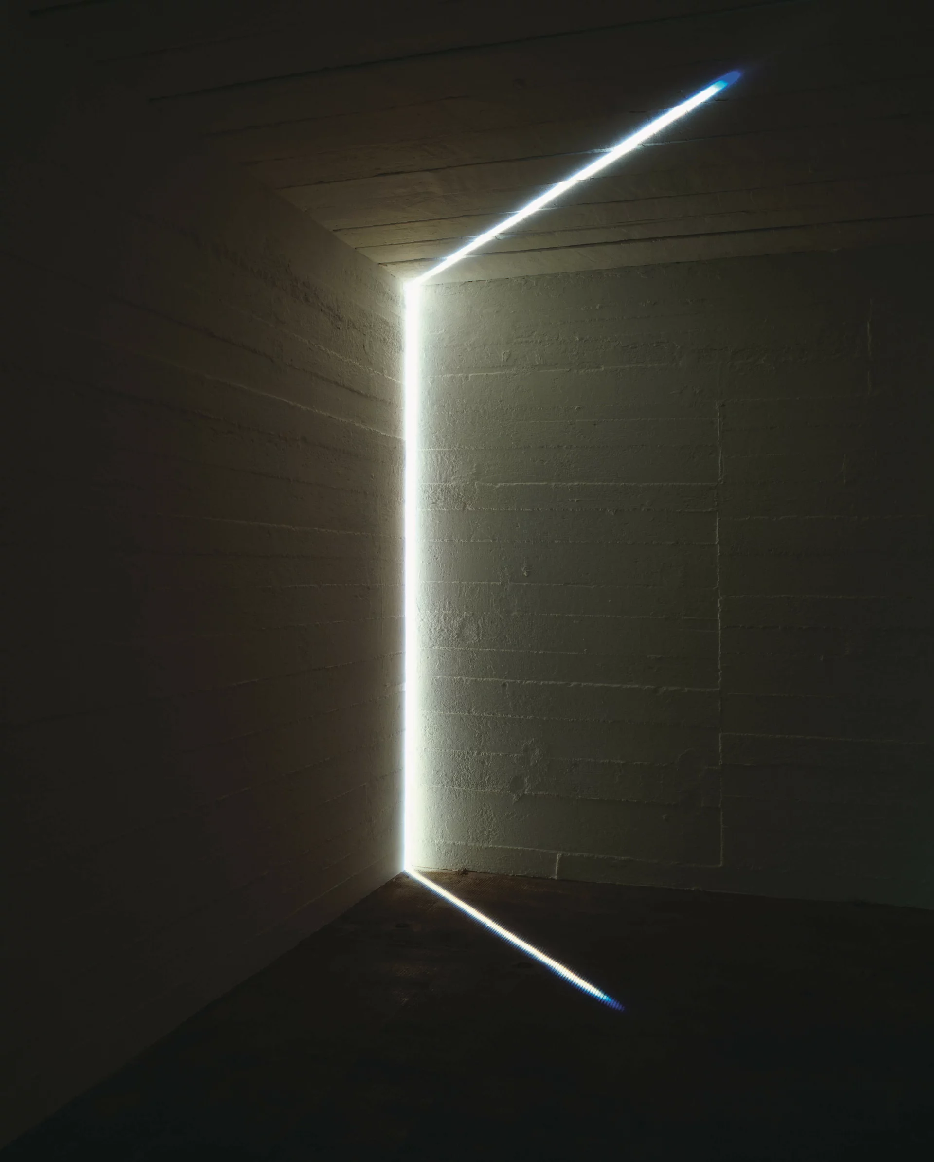 Feiner weißer Lichtstrahl durchzieht die Ecke eines dunklen Raumes und wirft geometrische Lichtlinien auf Wände, Boden und Decke.