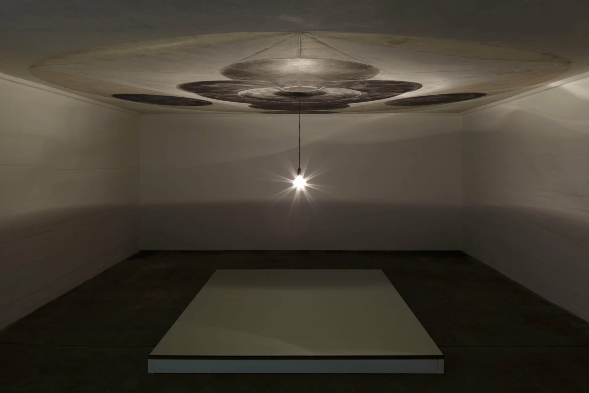 Installation von Dirk Bell mit einer einzelnen Glühbirne, die von der Decke hängt und eine großformatige, kreisförmige Zeichnung an der Decke beleuchtet; darunter eine erhöhte, helle Bodenfläche in einem dunklen Raum.