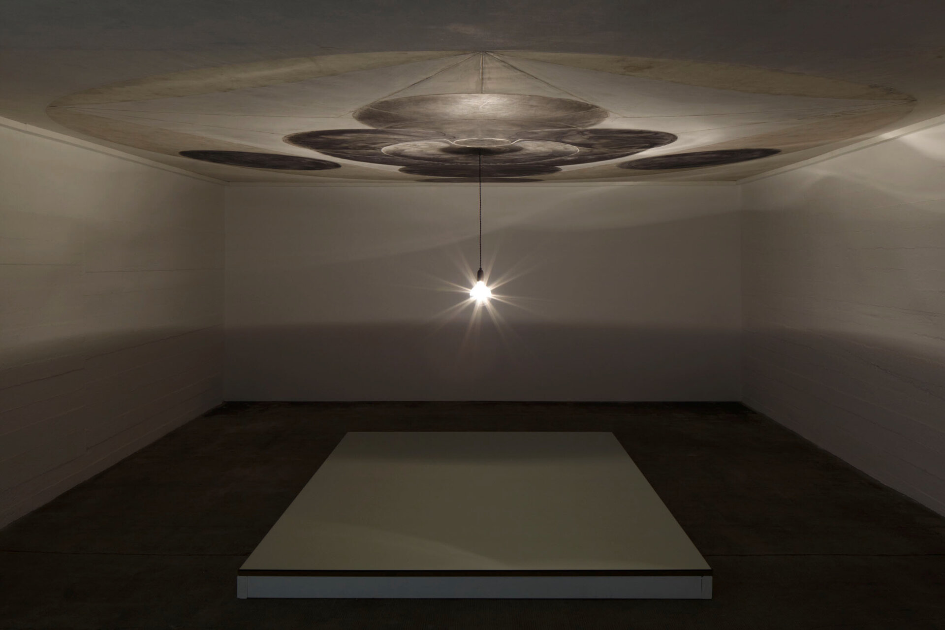 Installation von Dirk Bell mit einer einzelnen Glühbirne, die von der Decke hängt und eine großformatige, kreisförmige Zeichnung an der Decke beleuchtet; darunter eine erhöhte, helle Bodenfläche in einem dunklen Raum.
