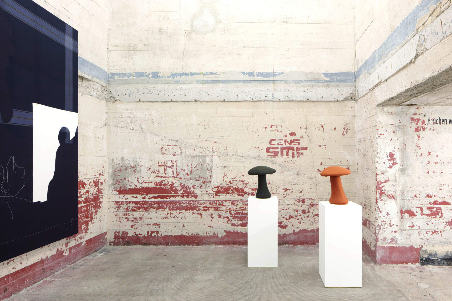 Zwei pilzartige Stoffskulpturen von Cosima von Bonin in Schwarz und Orange stehen auf weißen Sockeln in einem verwitterten Betonraum; links ein großformatiges textiles Wandbild in Dunkelblau und Weiß.