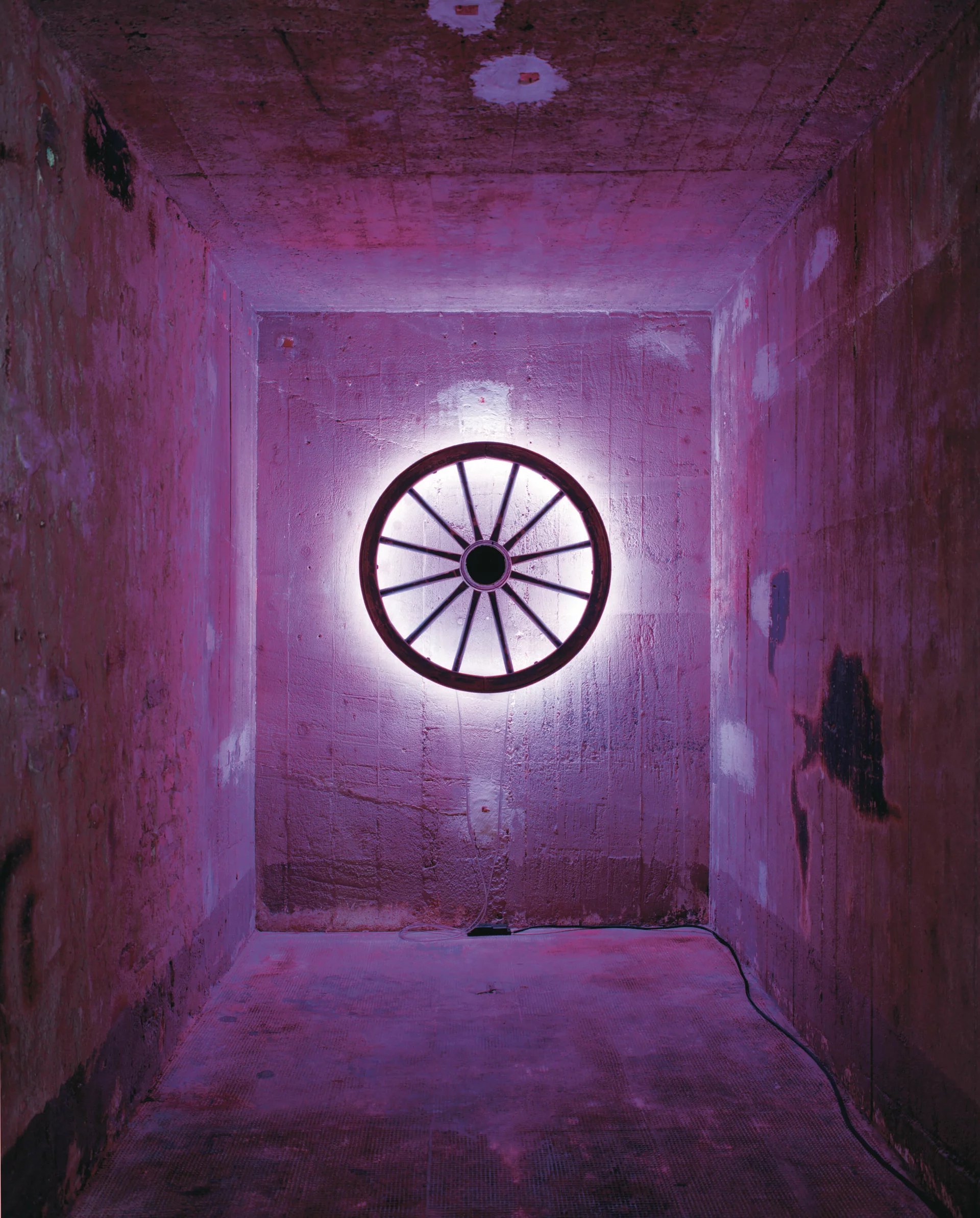 Ein beleuchtetes Wagenrad an einer Betonwand, umgeben von pink-violettem Licht in einem schmalen Raum.