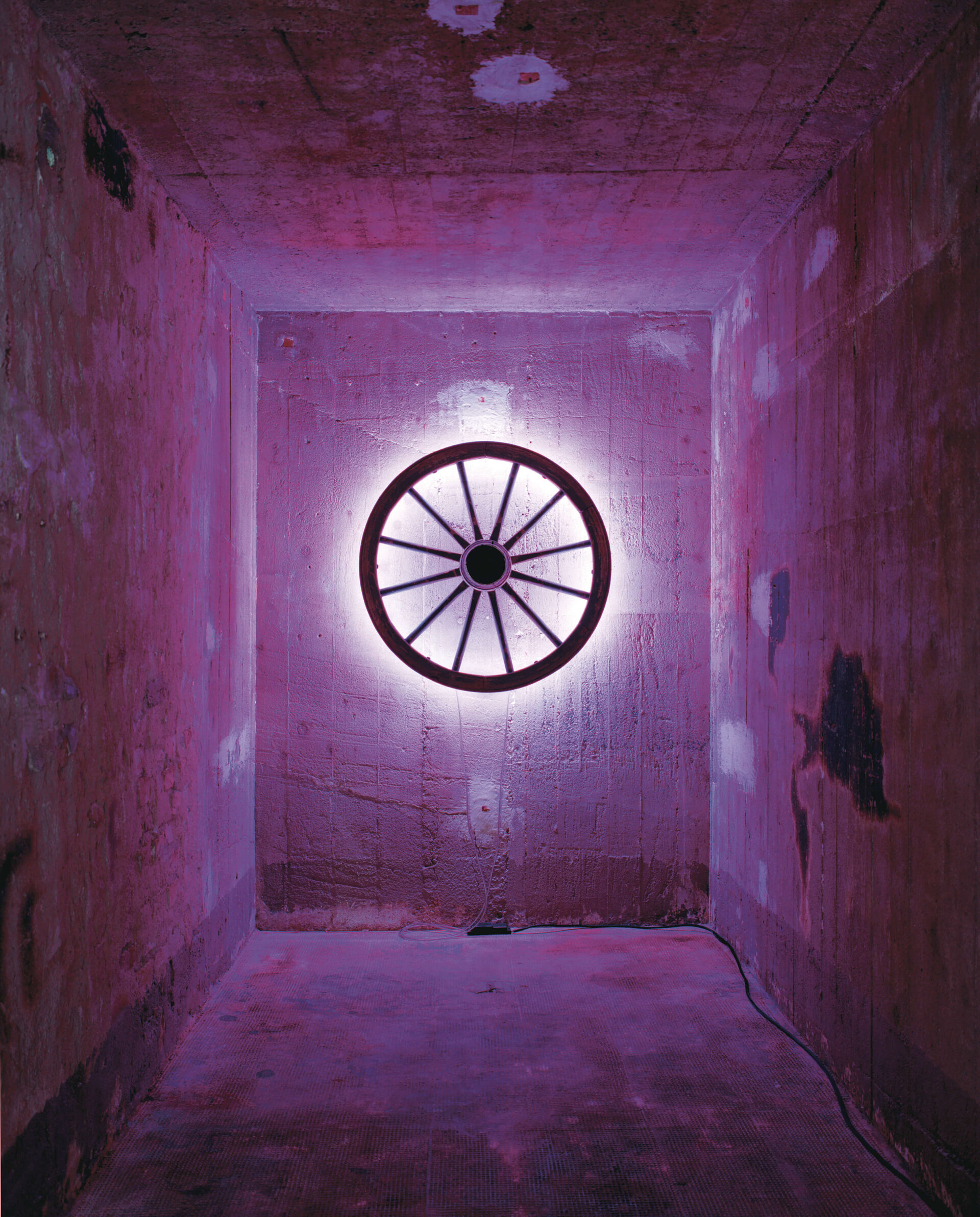 Ein beleuchtetes Wagenrad an einer Betonwand, umgeben von pink-violettem Licht in einem schmalen Raum.