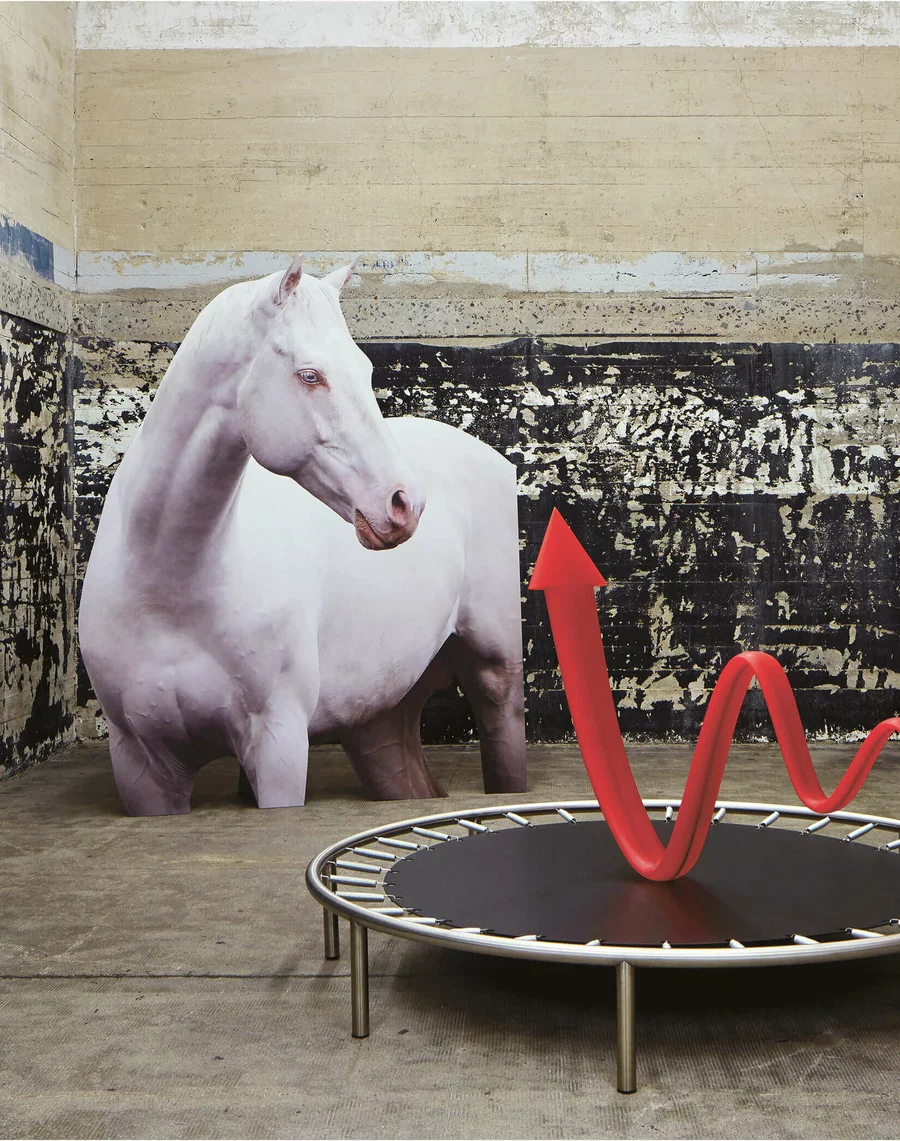 Installative Arbeit mit lebensgroßem Cut-out eines weißen Pferdes und einem roten, geschwungenen Pfeil, der aus einem Trampolin aufsteigt. Die Szene spielt vor einer stark verwitterten Wand im Boros Bunker Berlin.