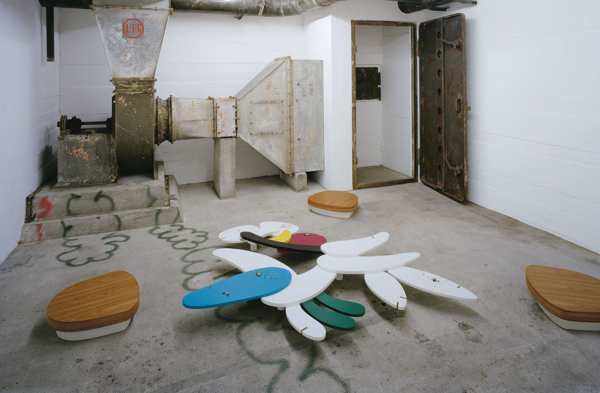 Bunte, blütenförmige Sitzskulpturen in industriellem Bunkerraum mit alten Belüftungsanlagen.