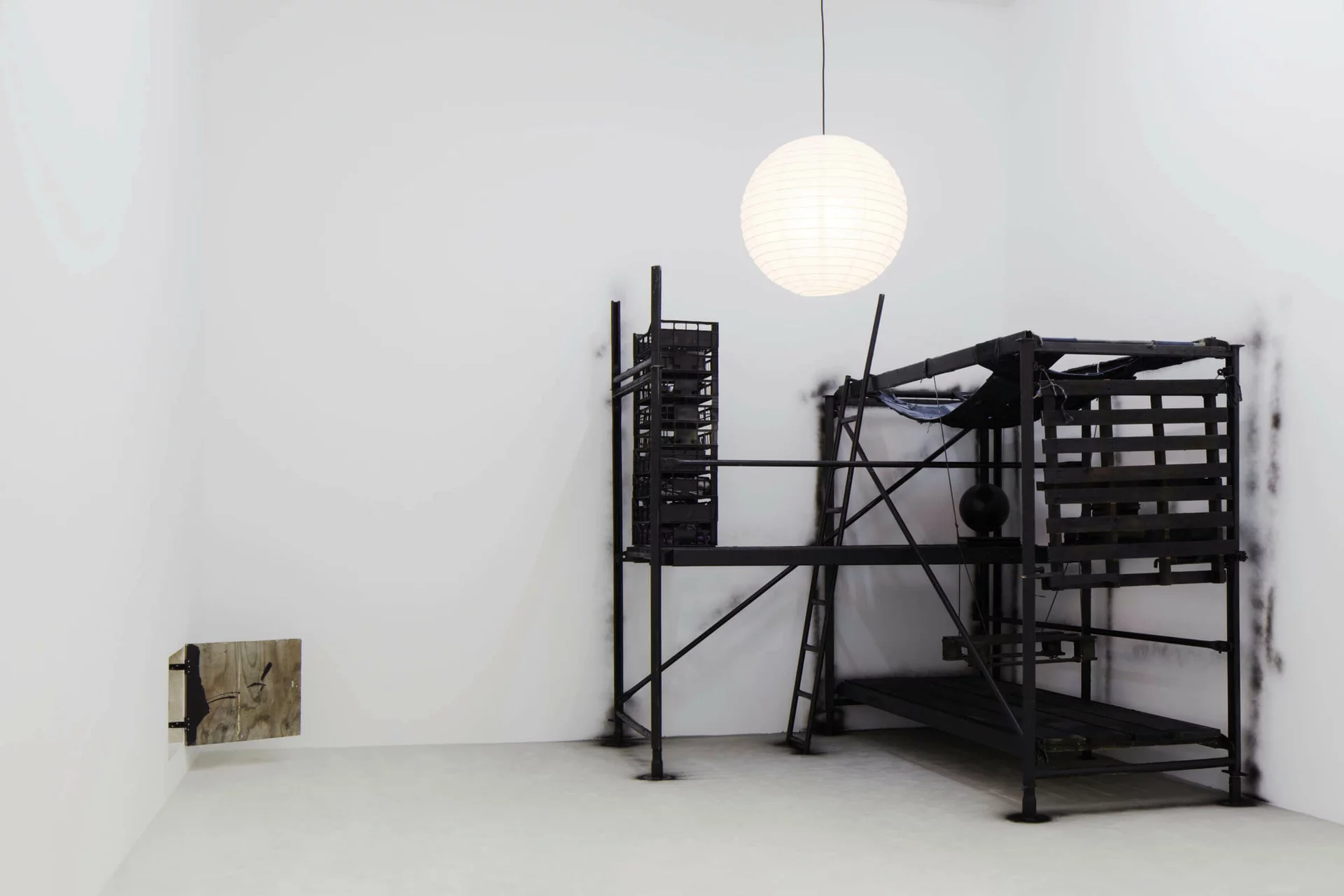 Installation von Klara Lidén mit einer schwarzen, grob gezimmerten Konstruktion aus Holzpaletten, Stahlstangen und einer Papierlampe; an der linken Wand eine aufklappbare Holzplatte mit Kritzeleien.